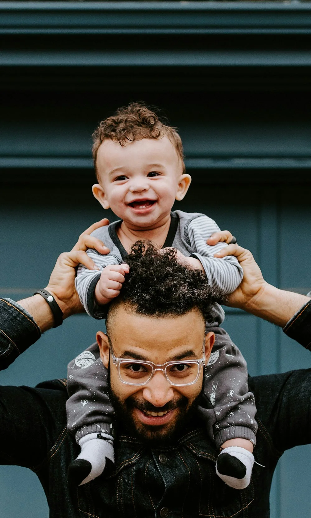A man with glasses and a beard is holding a young boy on his shoulders. The boy is smiling and wearing a striped shirt, and the man is smiling with a beard and glasses. They are both holding the boy's arms.