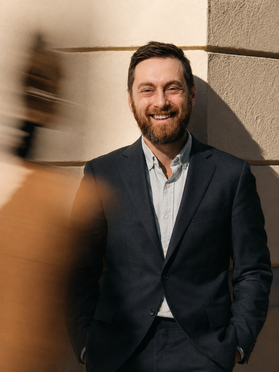 A man with a beard and short brown hair, smiling, wearing a dark suit jacket and a light-colored shirt, standing against a corner wall with sunlight casting shadows.