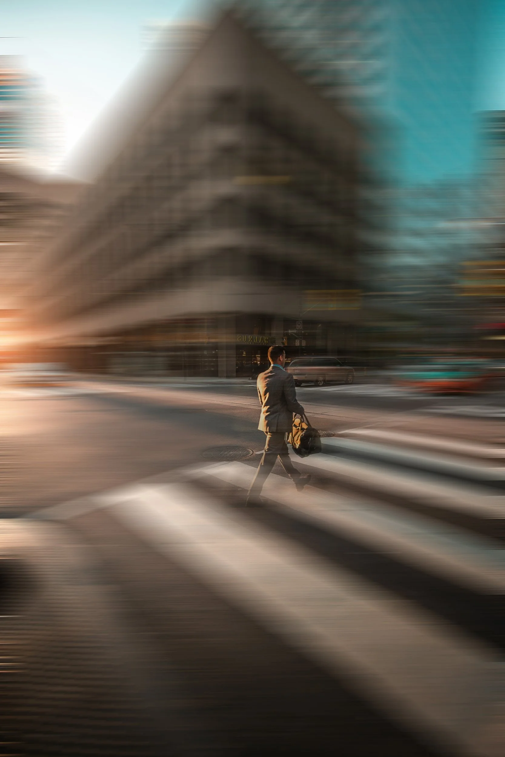 A man in a suit walking across a city street crosswalk with a rolling suitcase, with tall buildings in the background and bright sunlight.