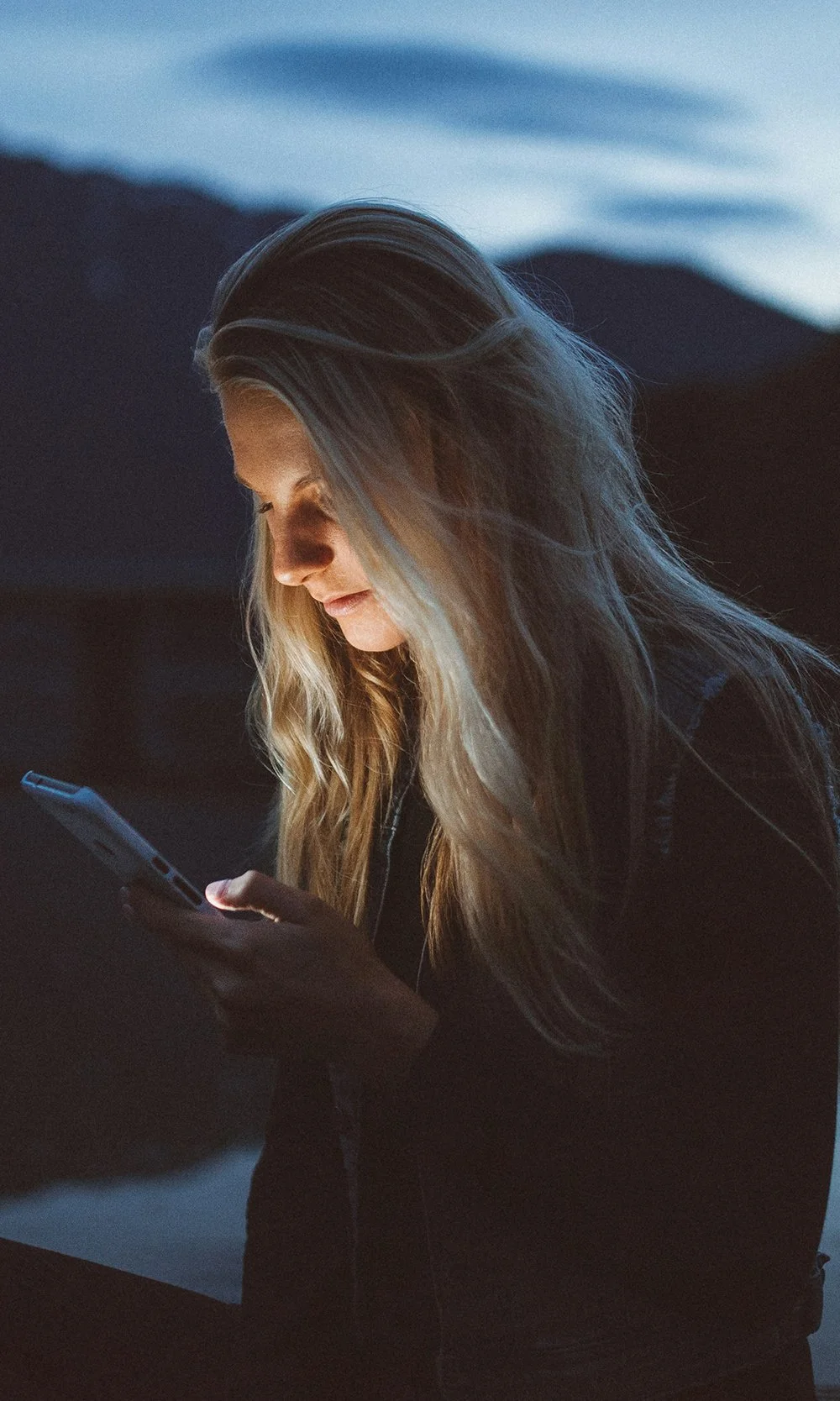 A woman with long blonde hair looking at her phone during dusk outdoors.