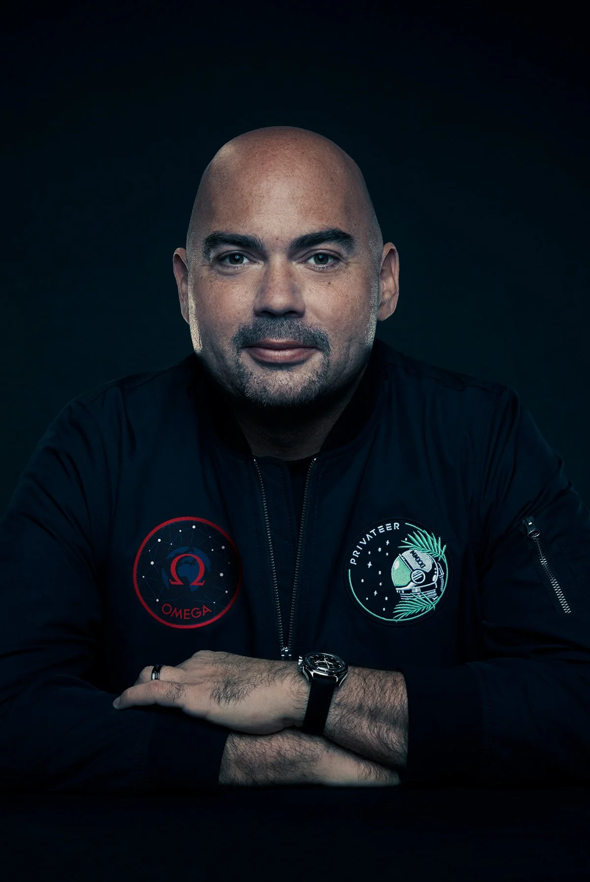 A man with a bald head and gray beard wearing a black jacket with patches, sitting with arms crossed against a black background.