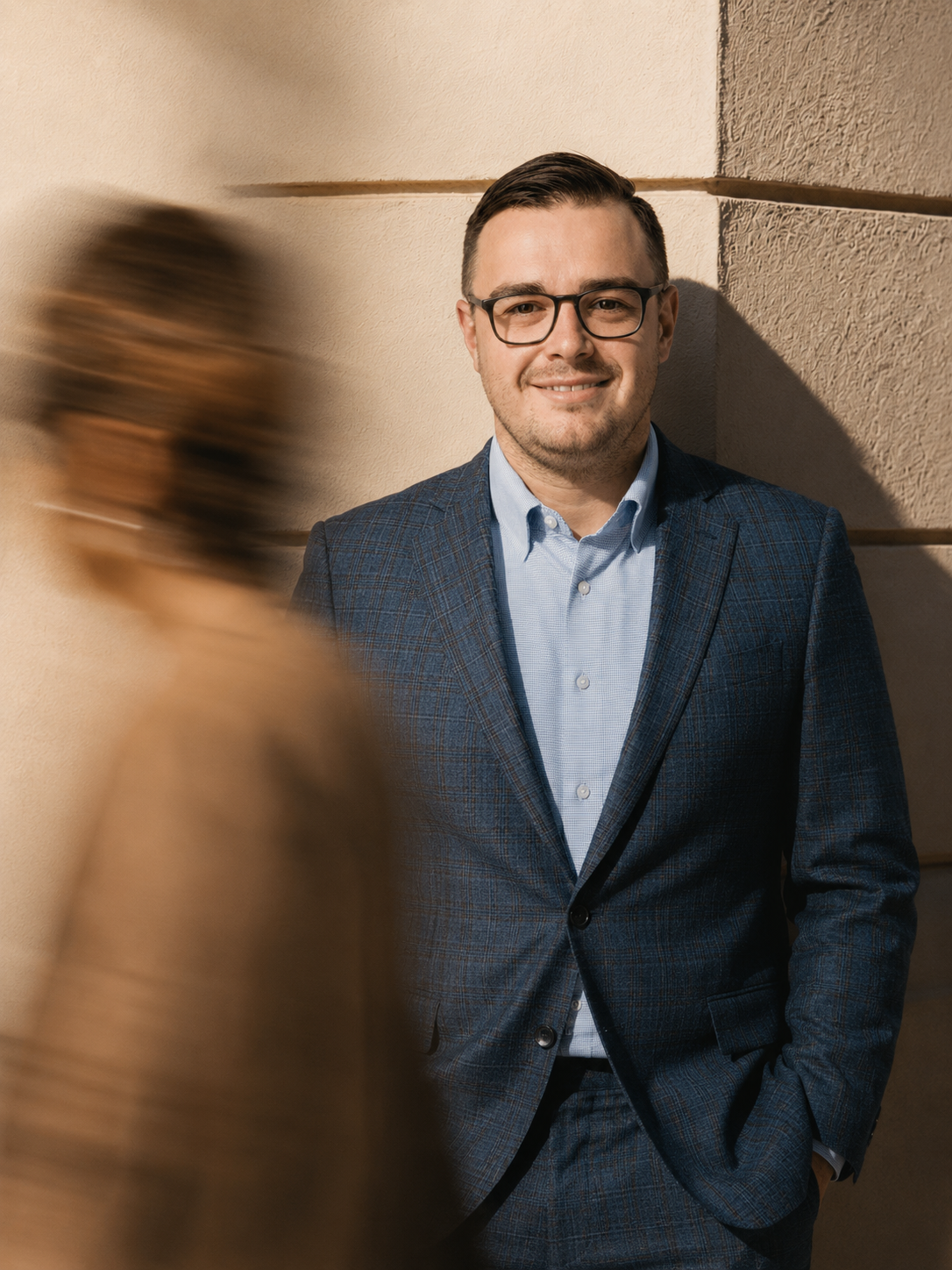 A man with dark hair, glasses, and a beard wearing a blue checkered blazer and light blue shirt, leaning against a beige and tan wall, smiling at the camera, with a blurred woman walking past in the foreground.