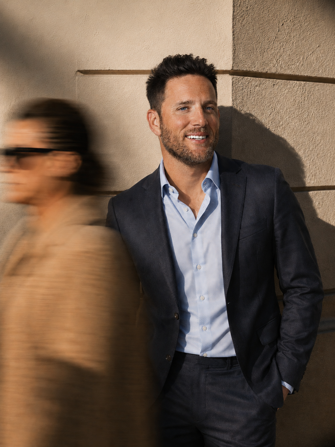 A man with brown hair and a beard leaning against a beige wall, smiling and wearing a dark blazer and light blue shirt. A woman with glasses and a tan coat is walking past him, blurred due to motion.
