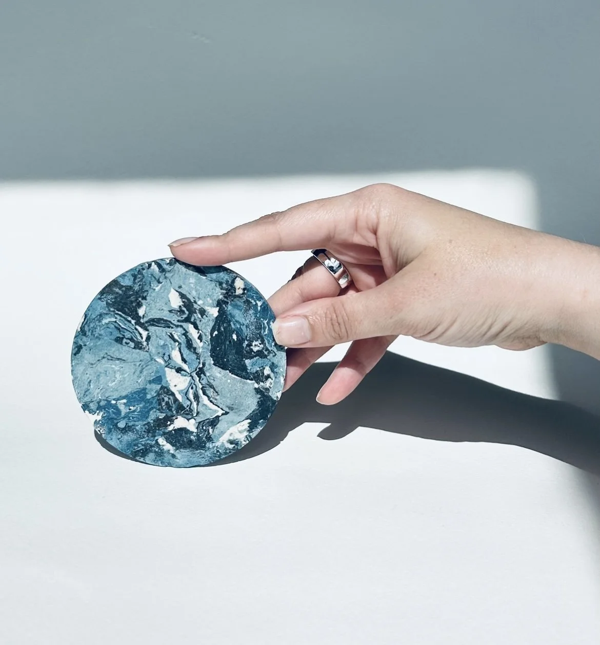 A hand with a silver ring on the finger holding a round piece of blue and white marble on a white surface.