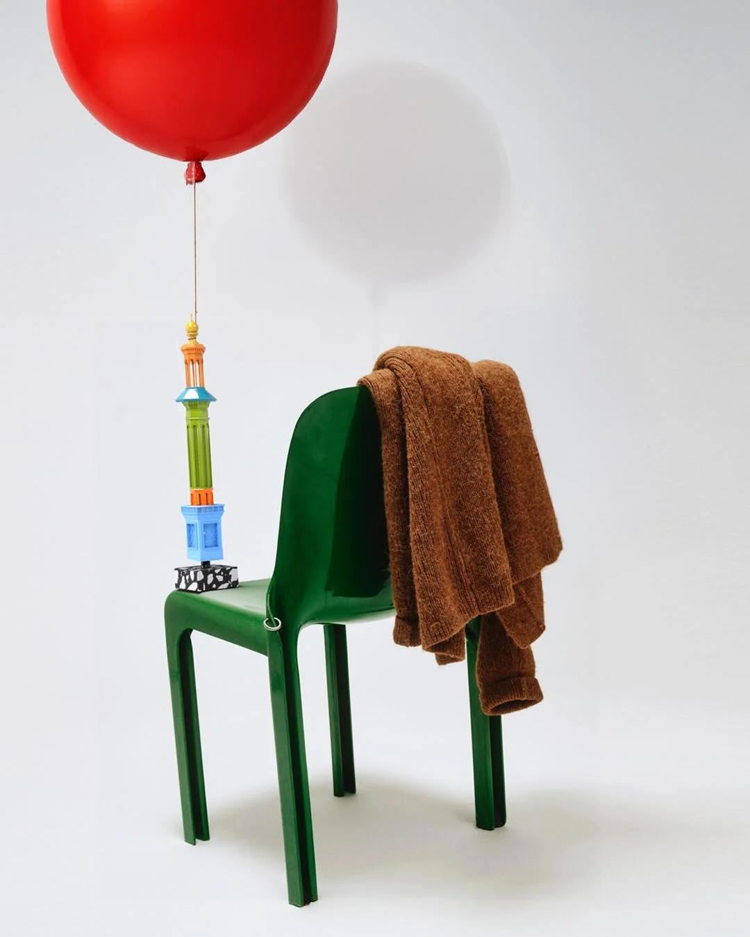 Green chair with a brown towel draped over it, a red balloon attached to the chair with a string, and a small toy tower hanging from the balloon's string, all set against a plain white background.