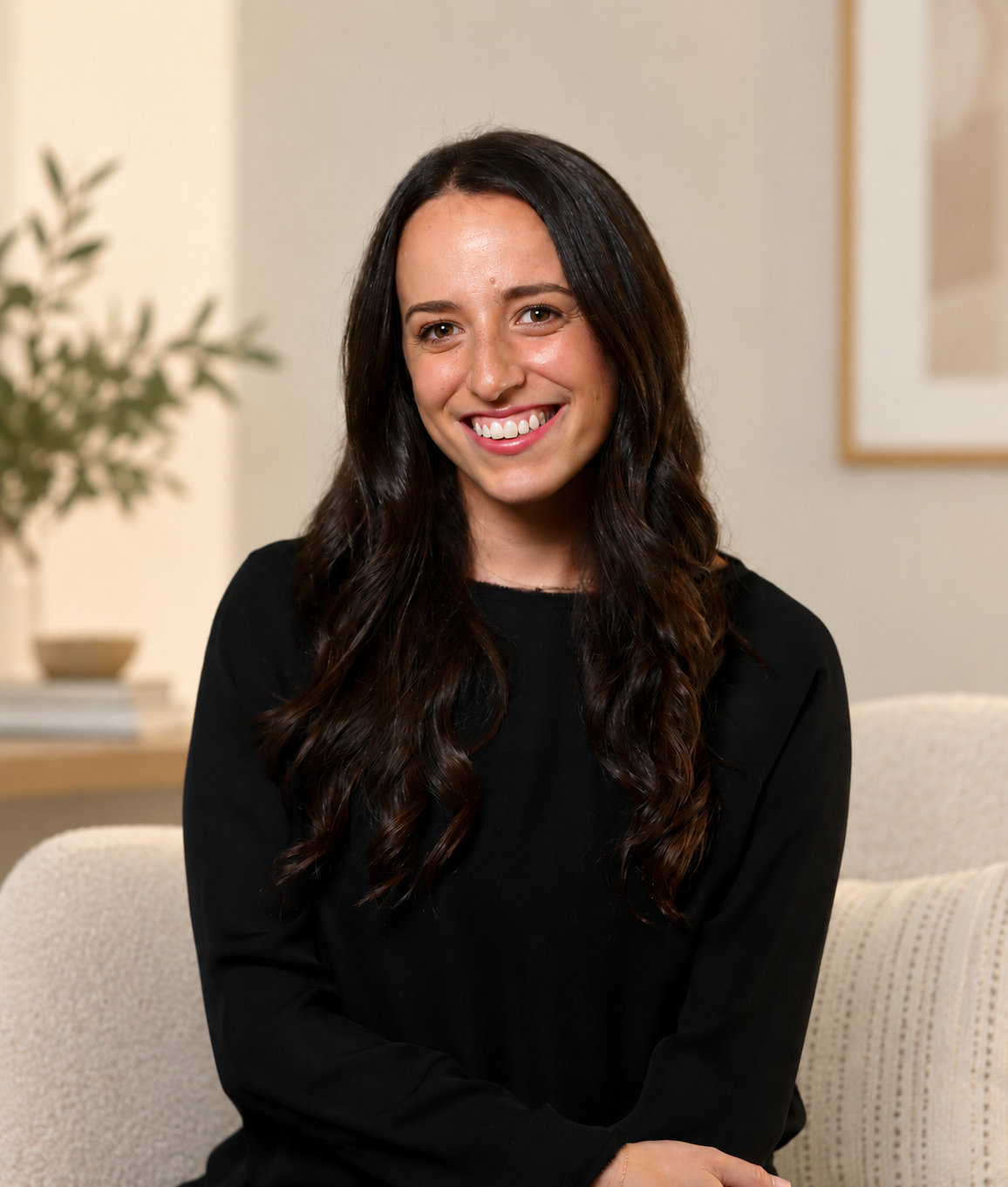 A woman with long, wavy dark hair, smiling, wearing a black top, sitting in a cozy, well-lit room with a plant and a framed picture on the wall behind her.