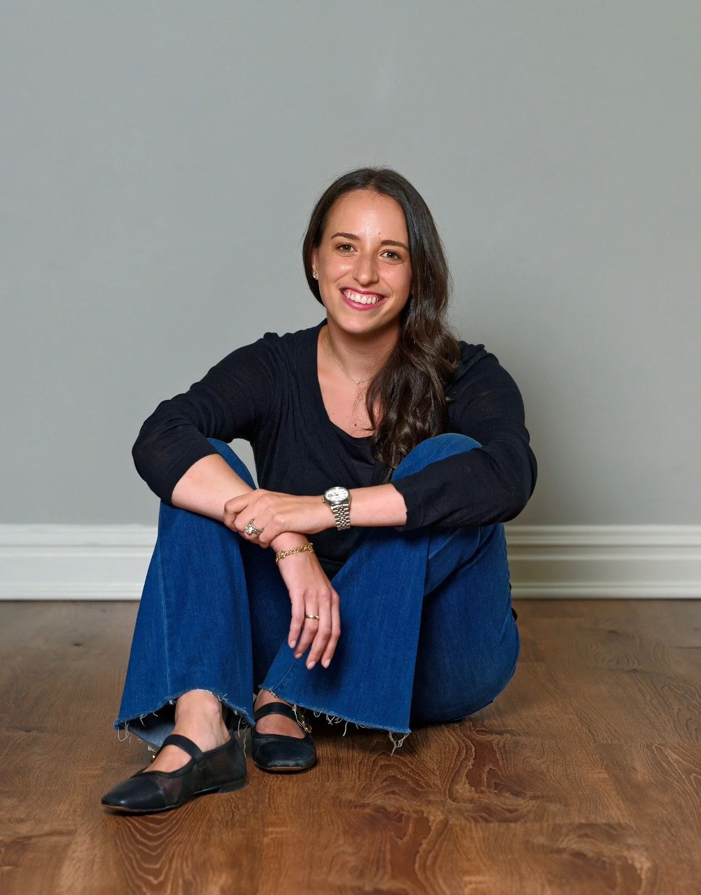 A woman with long brown hair, wearing a black long-sleeve shirt, blue jeans, black shoes, and jewelry, sitting on a wooden floor against a plain gray wall, smiling at the camera.