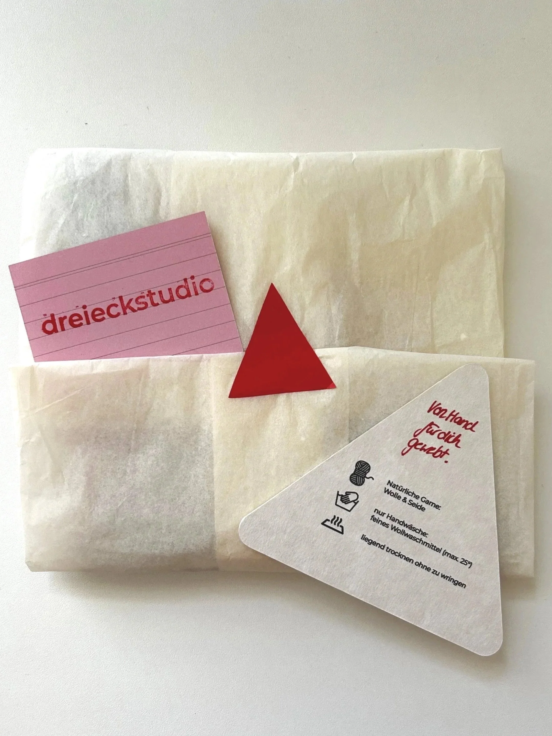 Eine Verpackung mit einem pinkfarbenen Zettel darauf, auf dem 'dreieckstudio' geschrieben steht, begleitet von einem weißen Zettel mit roten Worten und Symbolen für Waschanleitung.