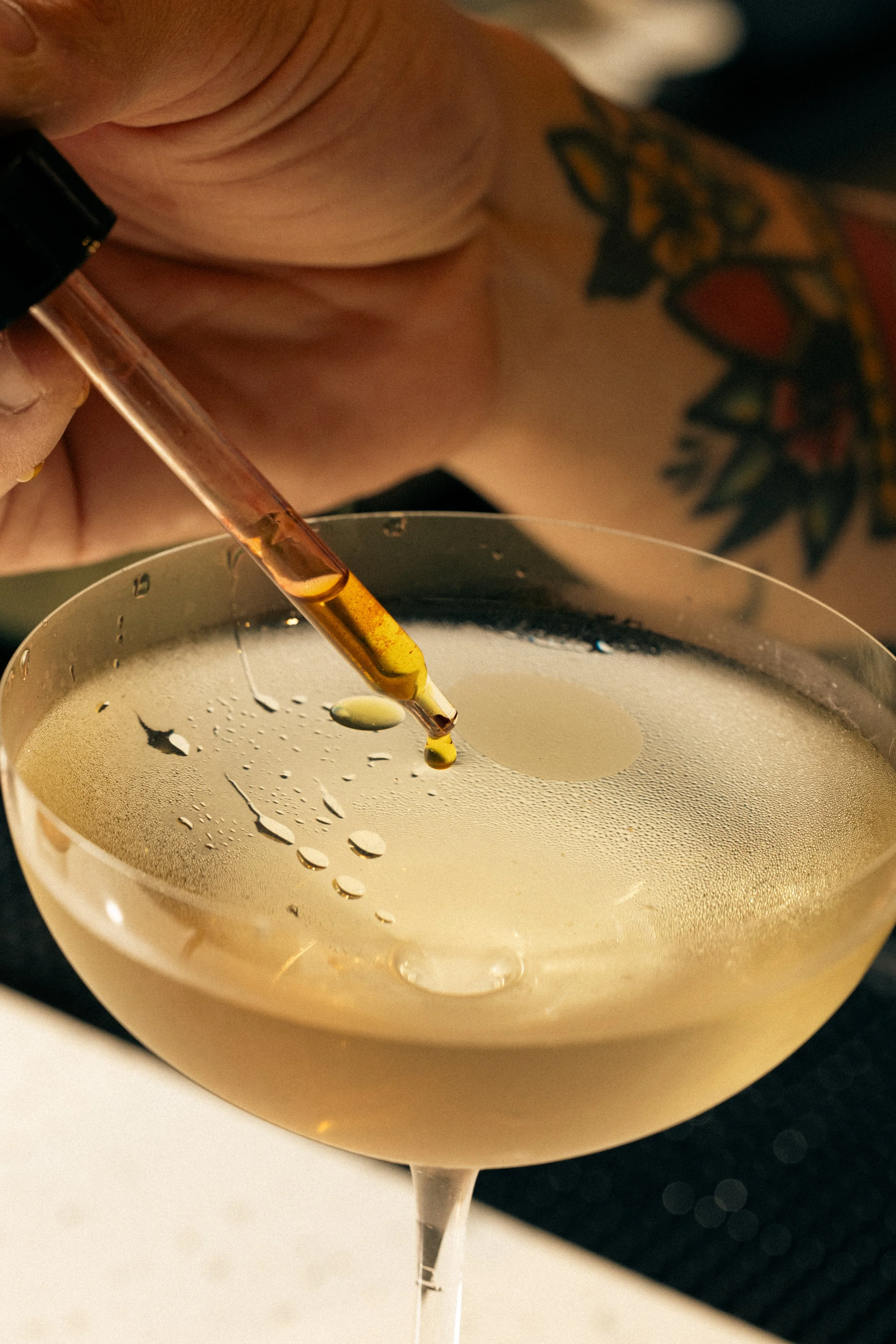 A cocktail glass filled with a creamy, light-colored beverage. A glass dropper is held above the drink, dispensing a few drops of a golden liquid.