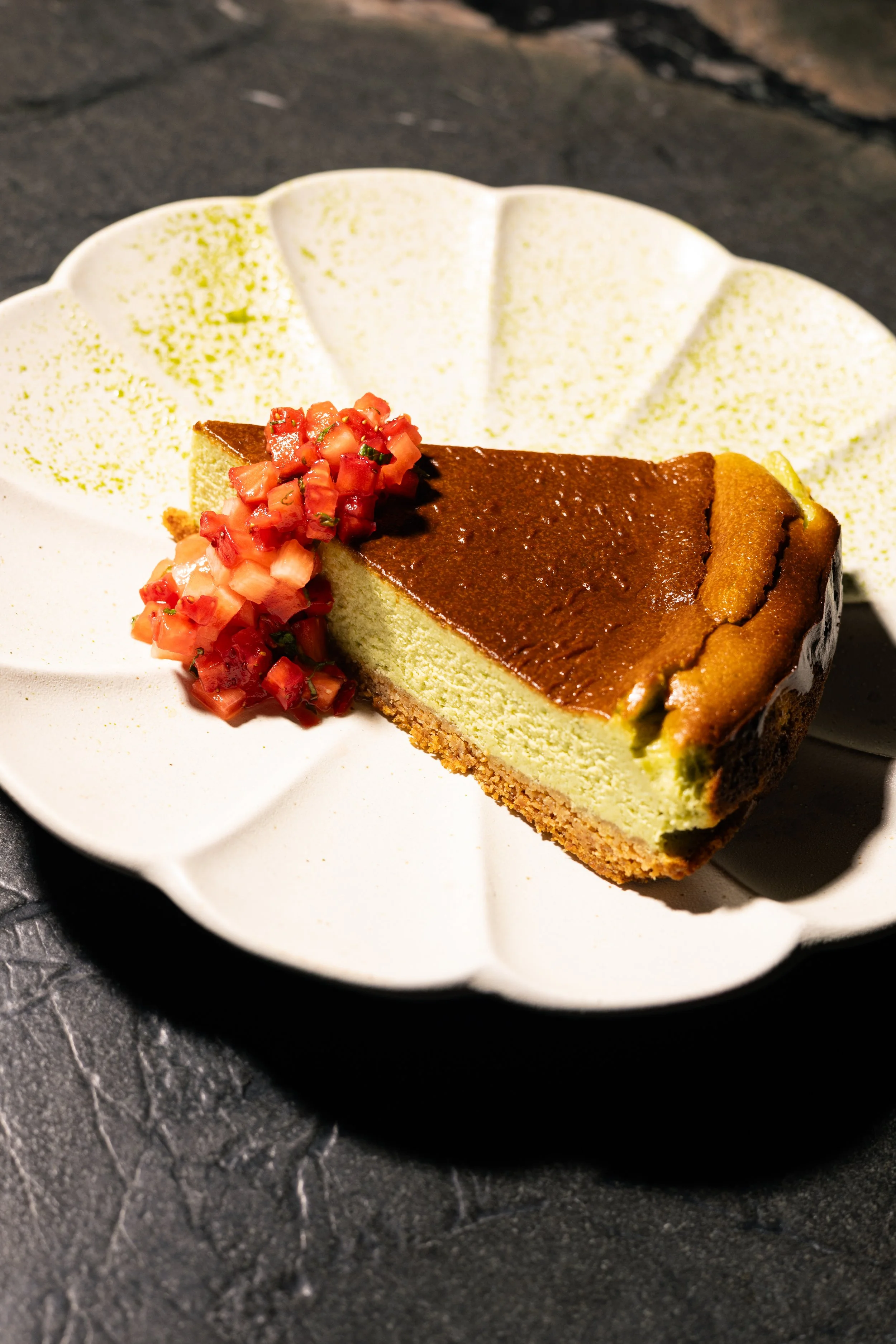 A slice of cheesecake topped with chopped strawberries and a serving of strawberry sauce, on a white decorative plate