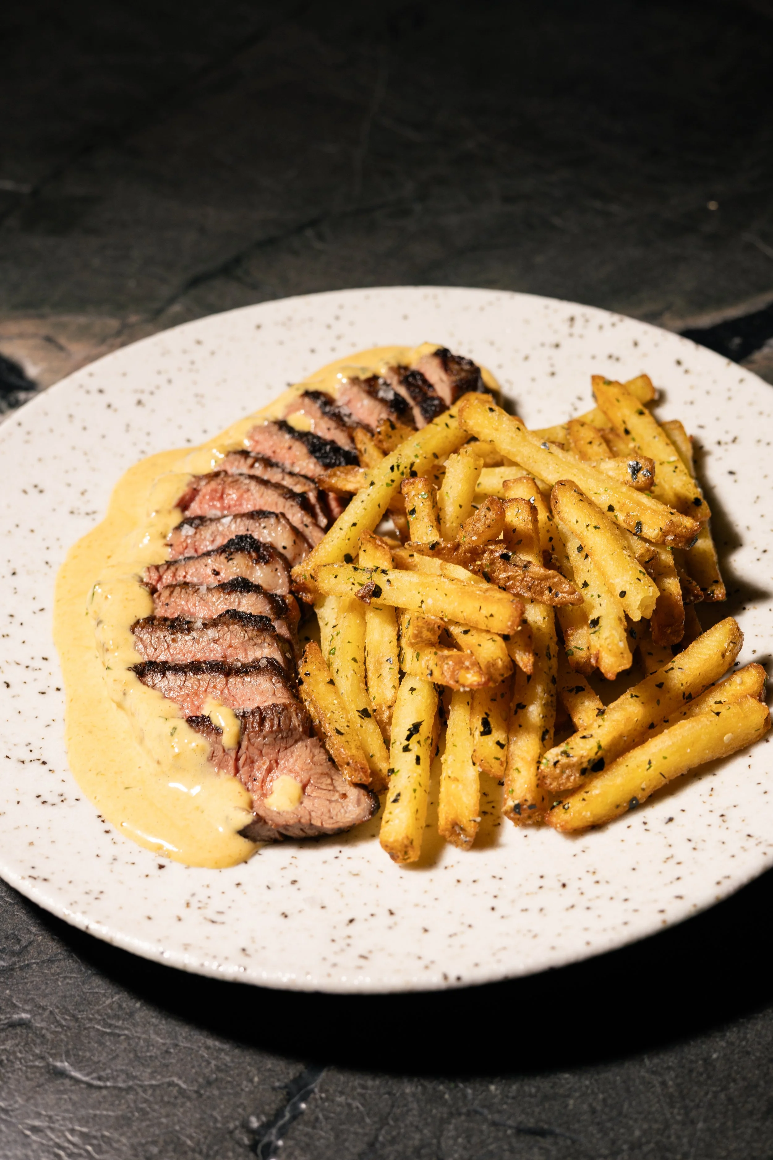 Sliced steak topped with a creamy sauce, served with French fries seasoned with herbs on a speckled white plate.
