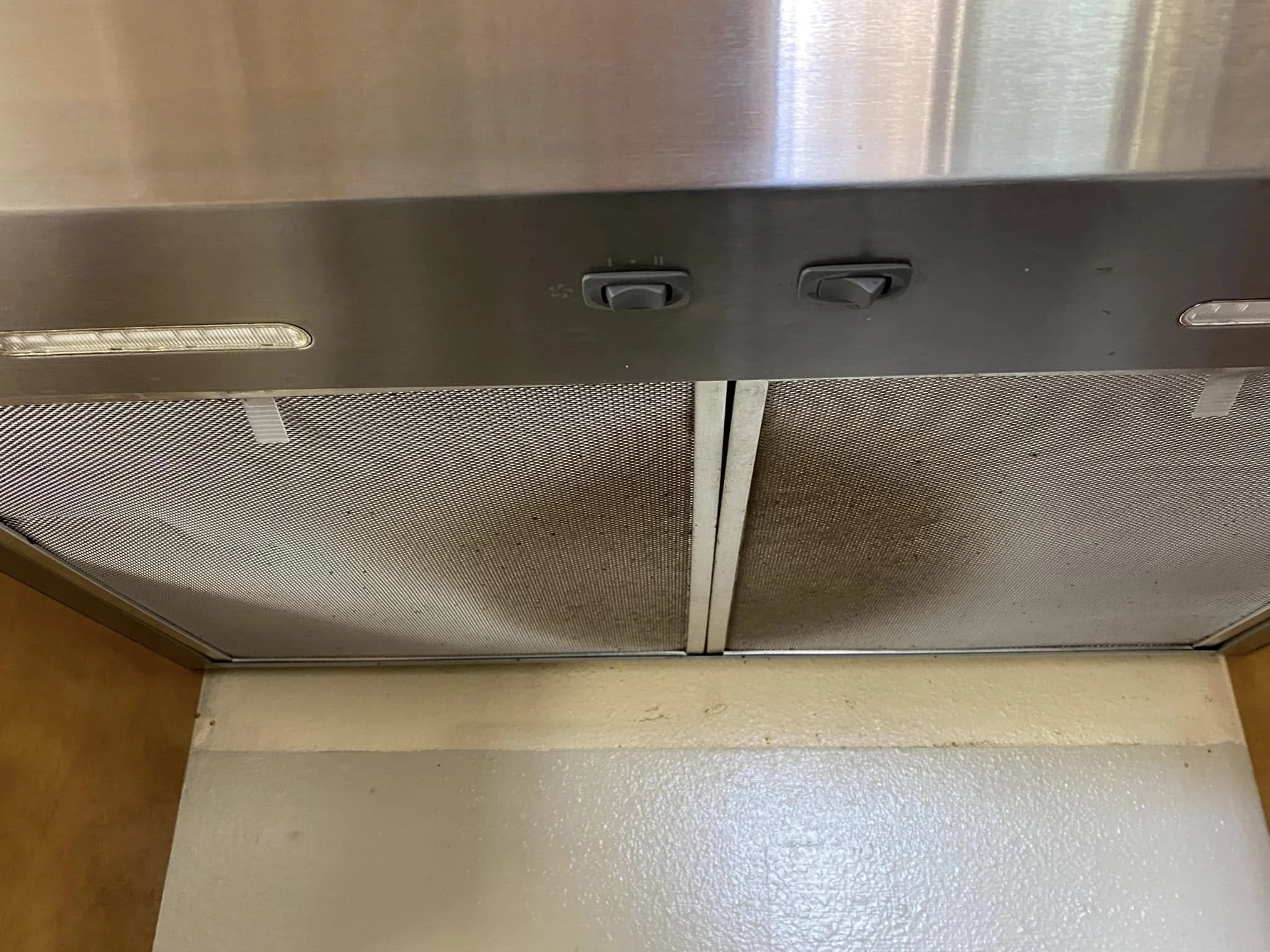 The image shows the underside of a stainless steel kitchen exhaust hood with two control switches, a light, and a vent, mounted above a beige textured wall and a beige floor.