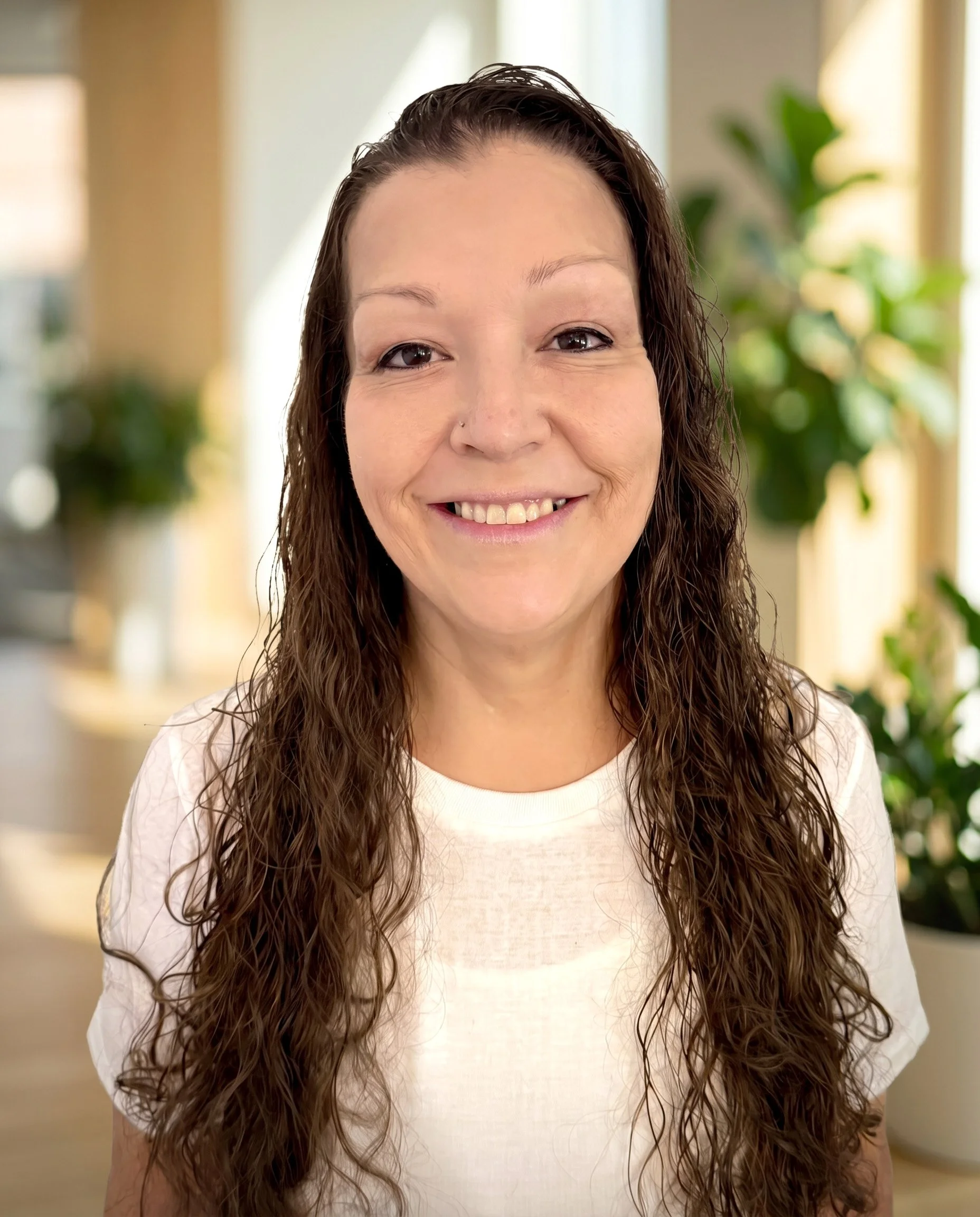 Close-up of a smiling woman with long, wavy brown hair, wearing a white shirt, standing indoors with blurred green plants in the background.