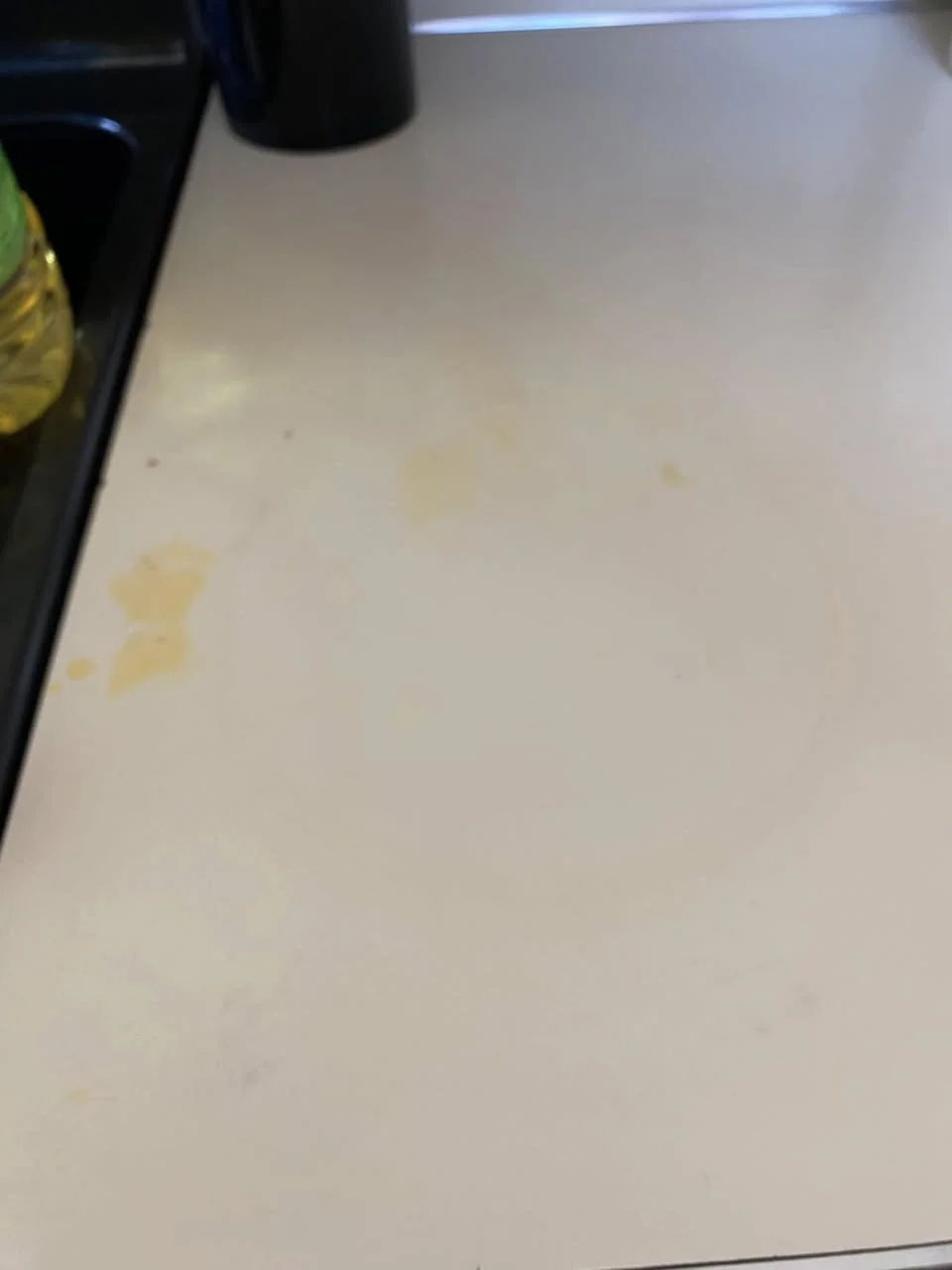 A beige countertop with some yellow stains, a black coffee mug, and a partial view of a black stovetop with a yellow bottle on it.