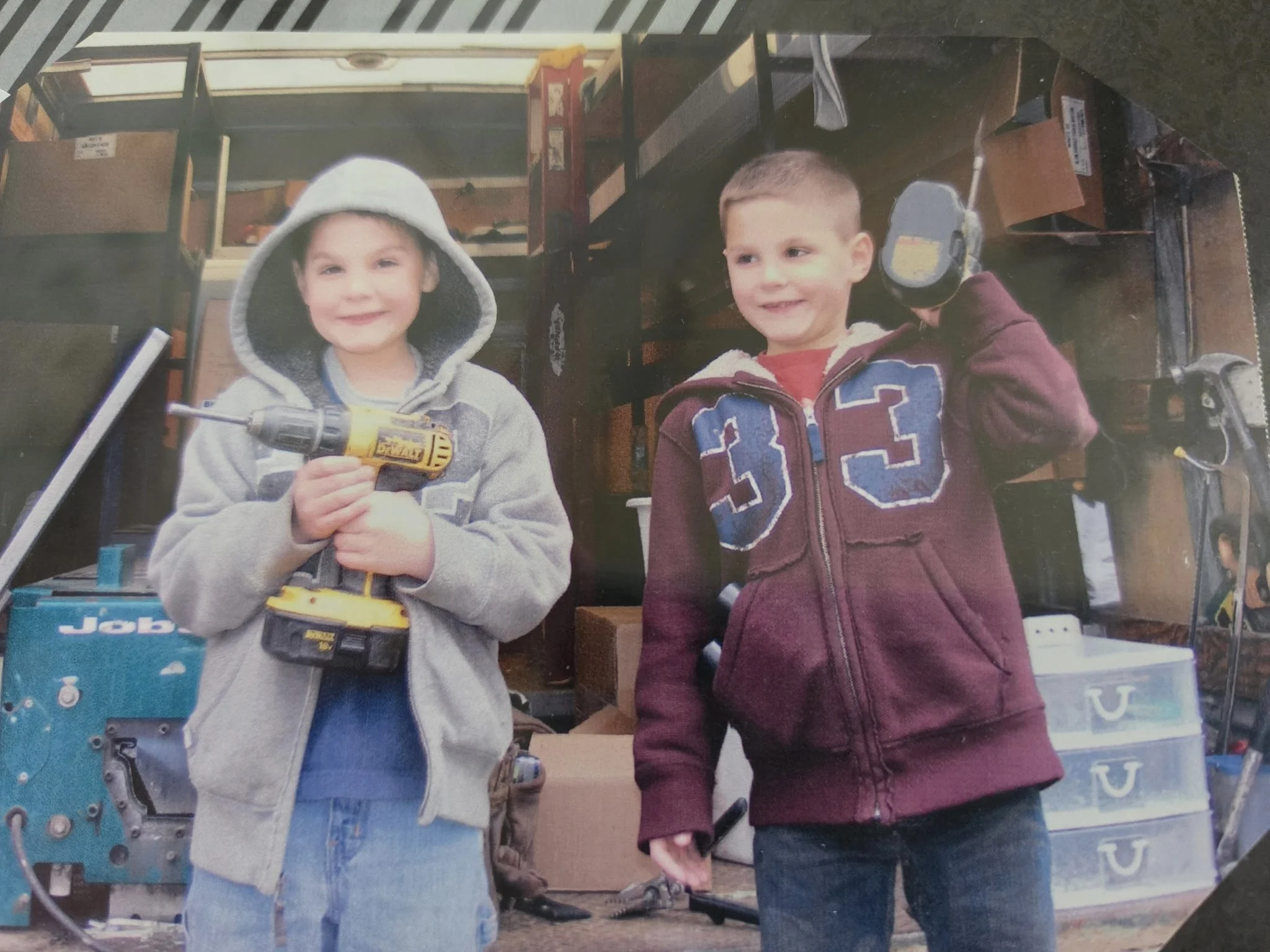 Two young boys smiling and holding hands in front of a toy store display. One boy is holding a yellow power drill. The other boy is wearing a maroon hoodie with the number 23.
