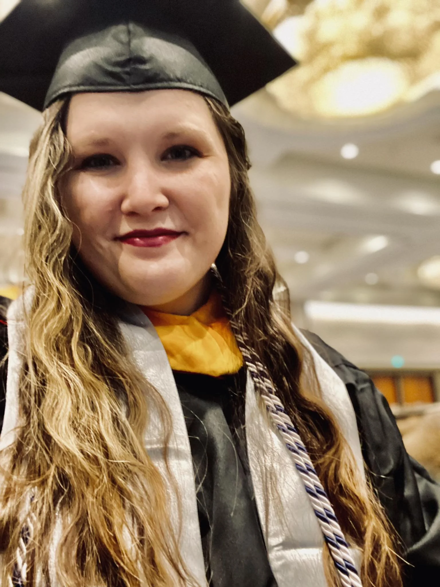 Close-up of a woman in a graduation cap and gown smiling indoors with a fancy ceiling in the background.
