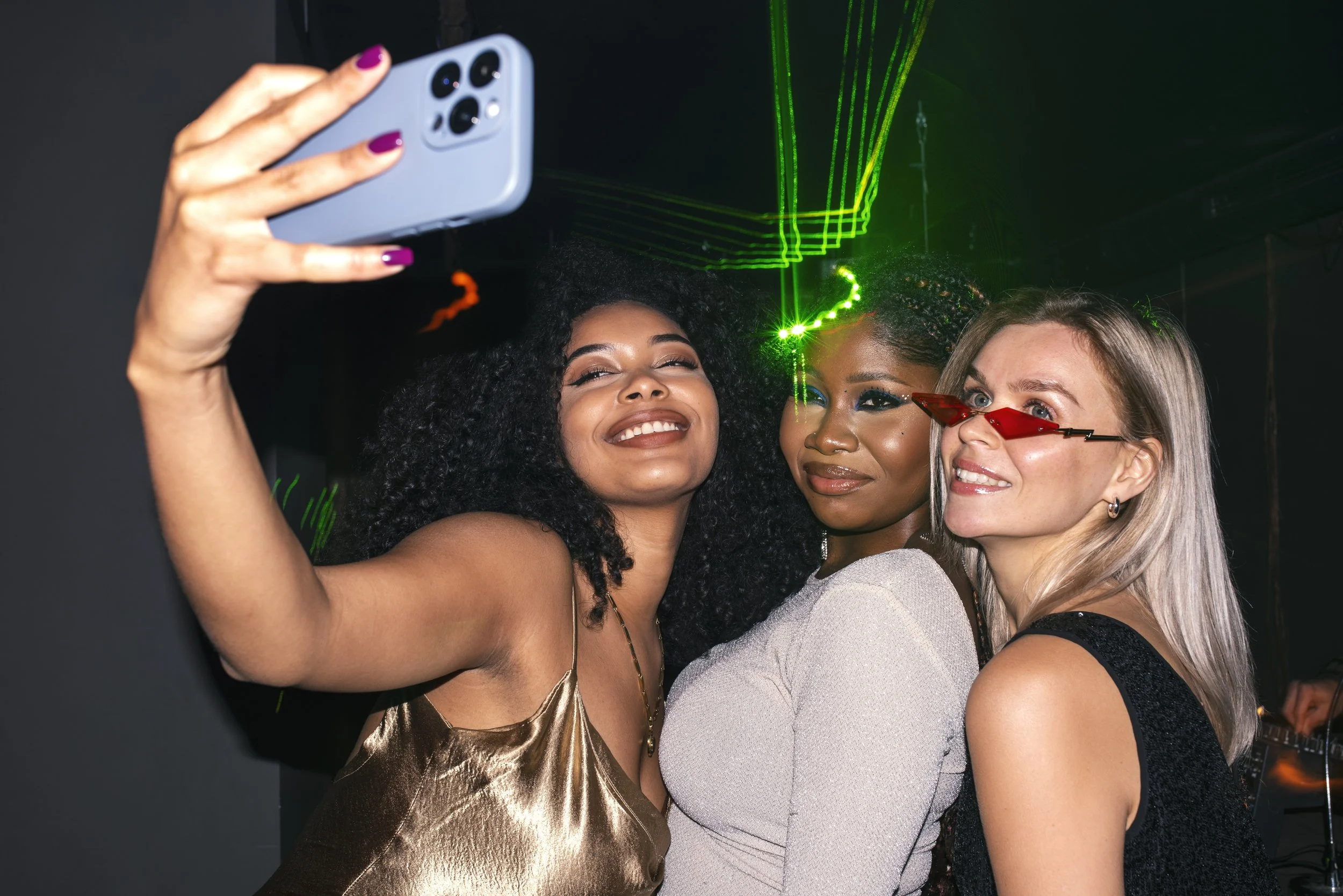 Three women taking a selfie at a party with green laser lights in the background.