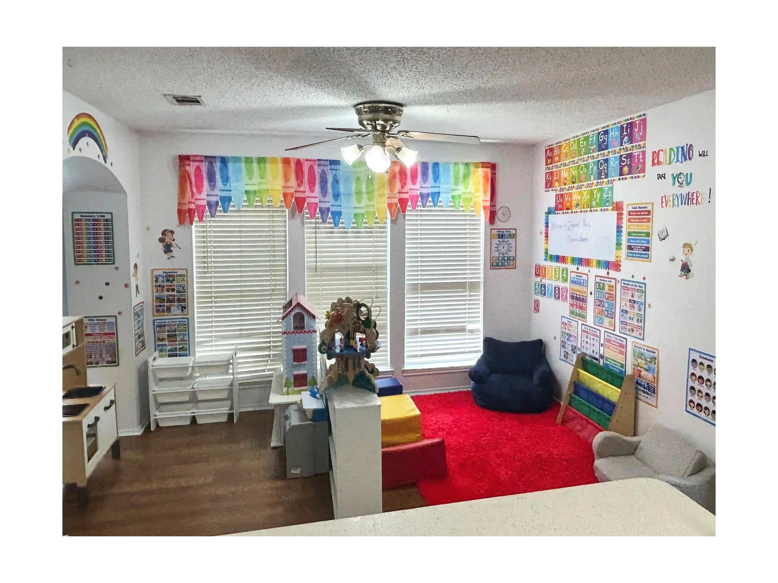 A colorful classroom corner featuring rainbow-themed decorations, educational posters related to the alphabet, numbers, and reading, along with a whiteboard, a small sofa, plush toys, and a red rug.