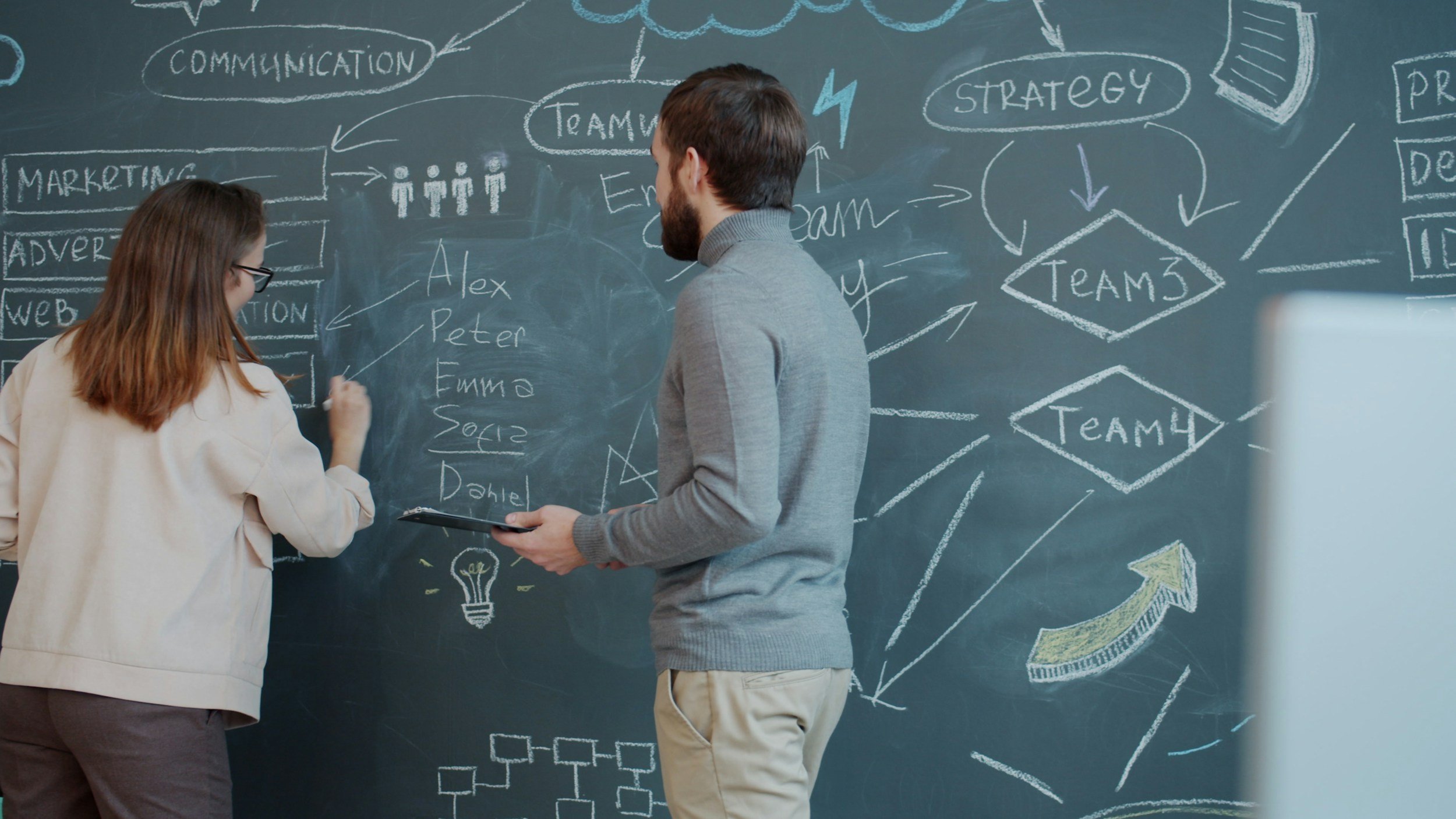 Two people, a woman and a man, are standing in front of a chalkboard filled with diagrams and words related to teamwork, strategy, and communication. The woman is writing on the board while the man is holding a tablet.