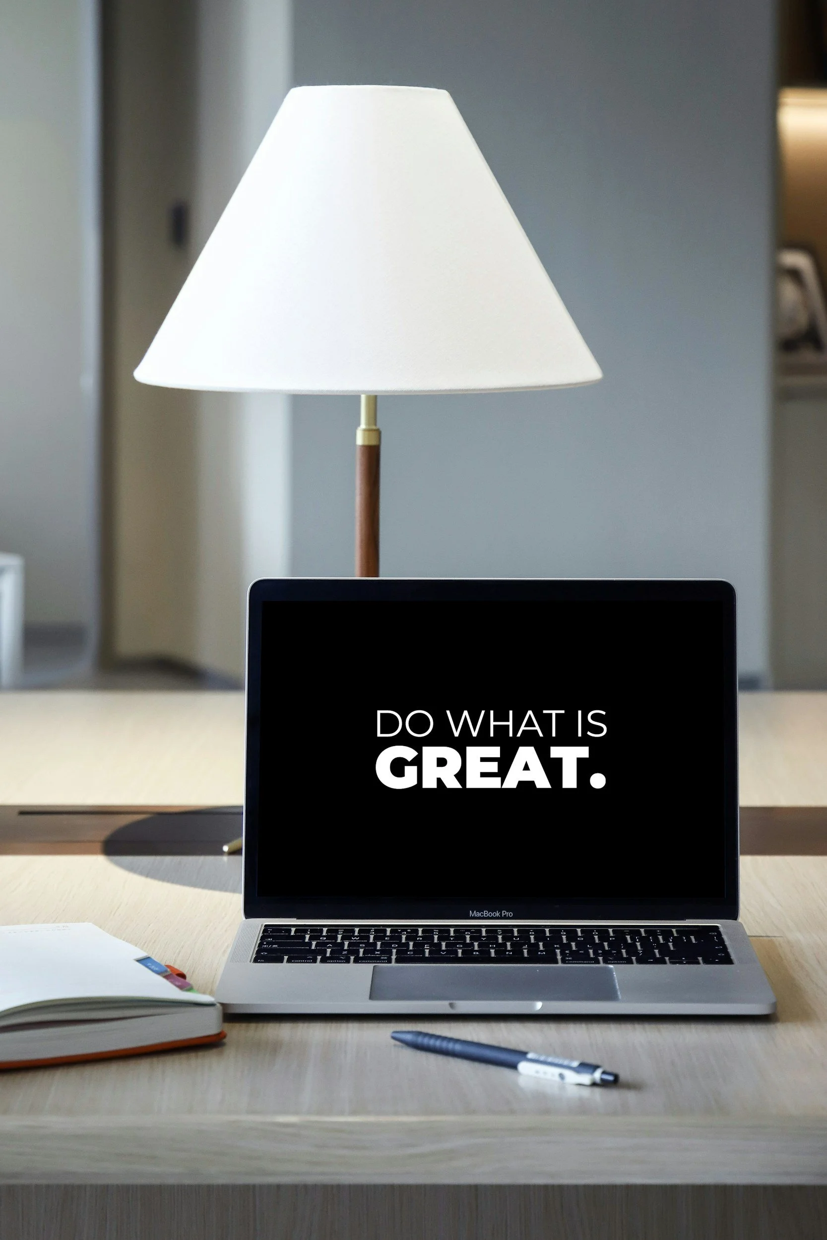 A computer screen displaying the text 'Do what is great.' on a black background, placed on a wooden desk with a pen, open notebook, and a closed laptop, with a lamp behind it in a modern office or home workspace.