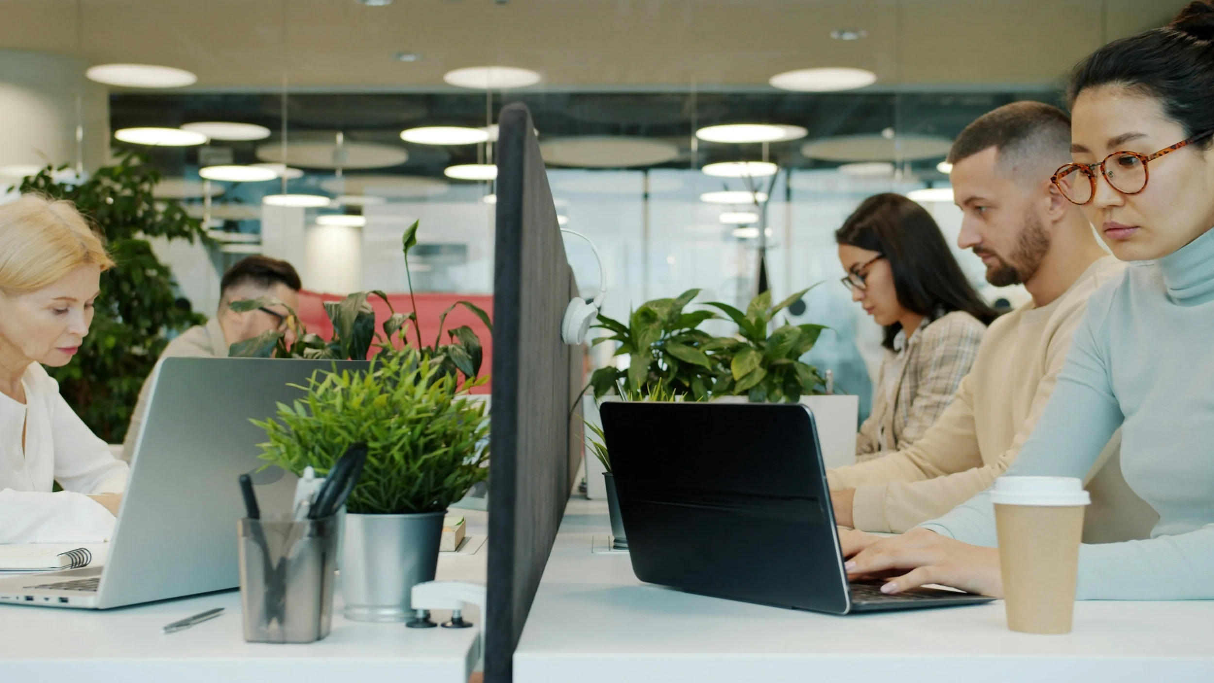 A group of diverse people working at their desks in an open office environment with plants and computers.