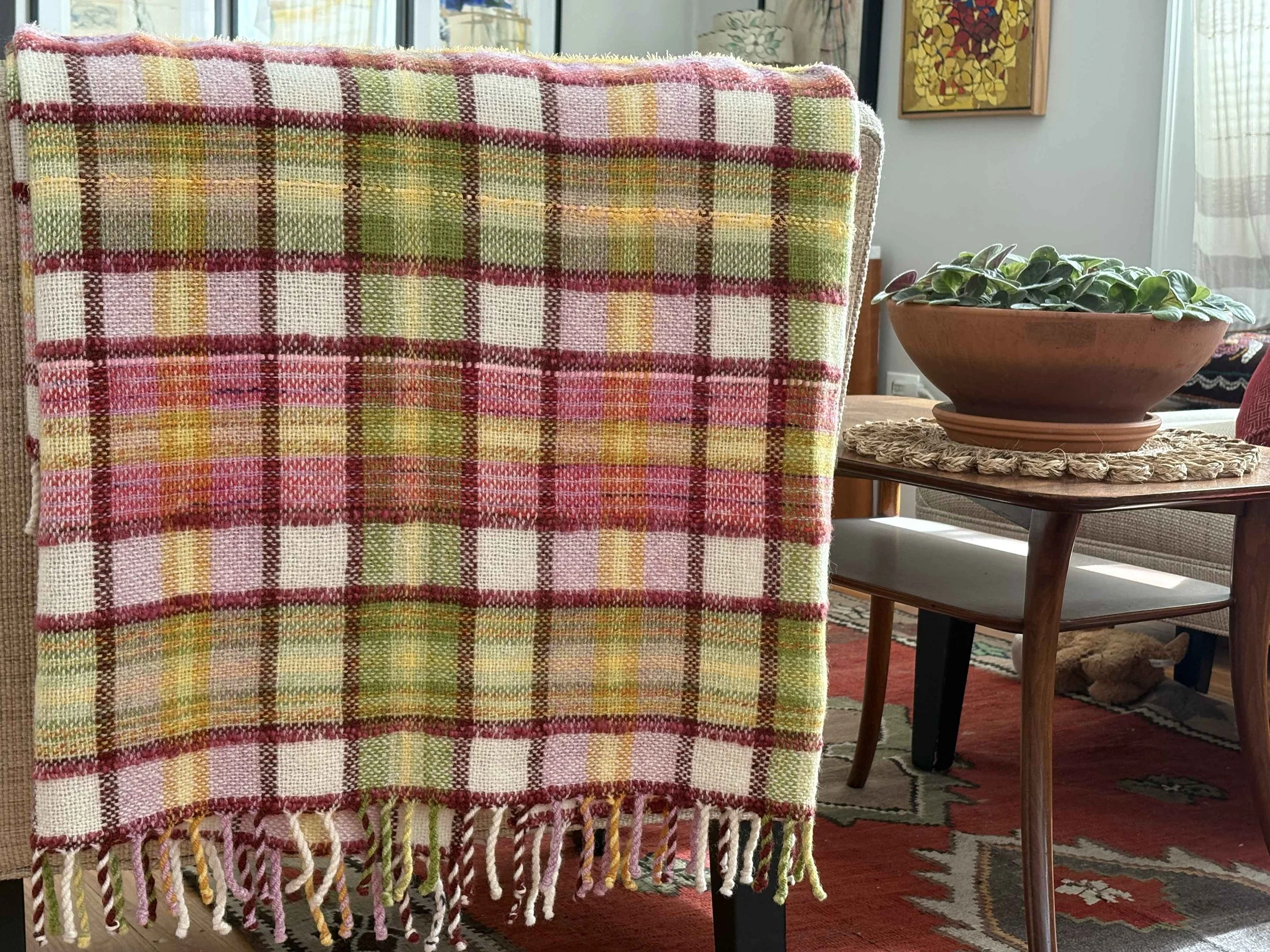 A colorful checkered woven blanket with fringes draped over the back of a chair, next to a round terracotta planter with a lush green plant on a wooden table. In the background, there is a window with curtains, a piece of wall art, and part of a patterned rug visible.