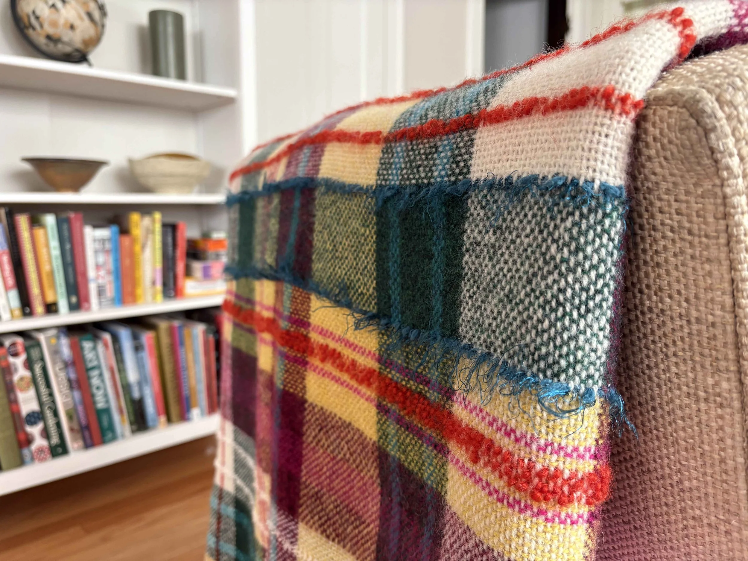 Close-up of a folded colorful plaid blanket draped over a textured beige armrest, with a bookshelf and decorative objects in the background.