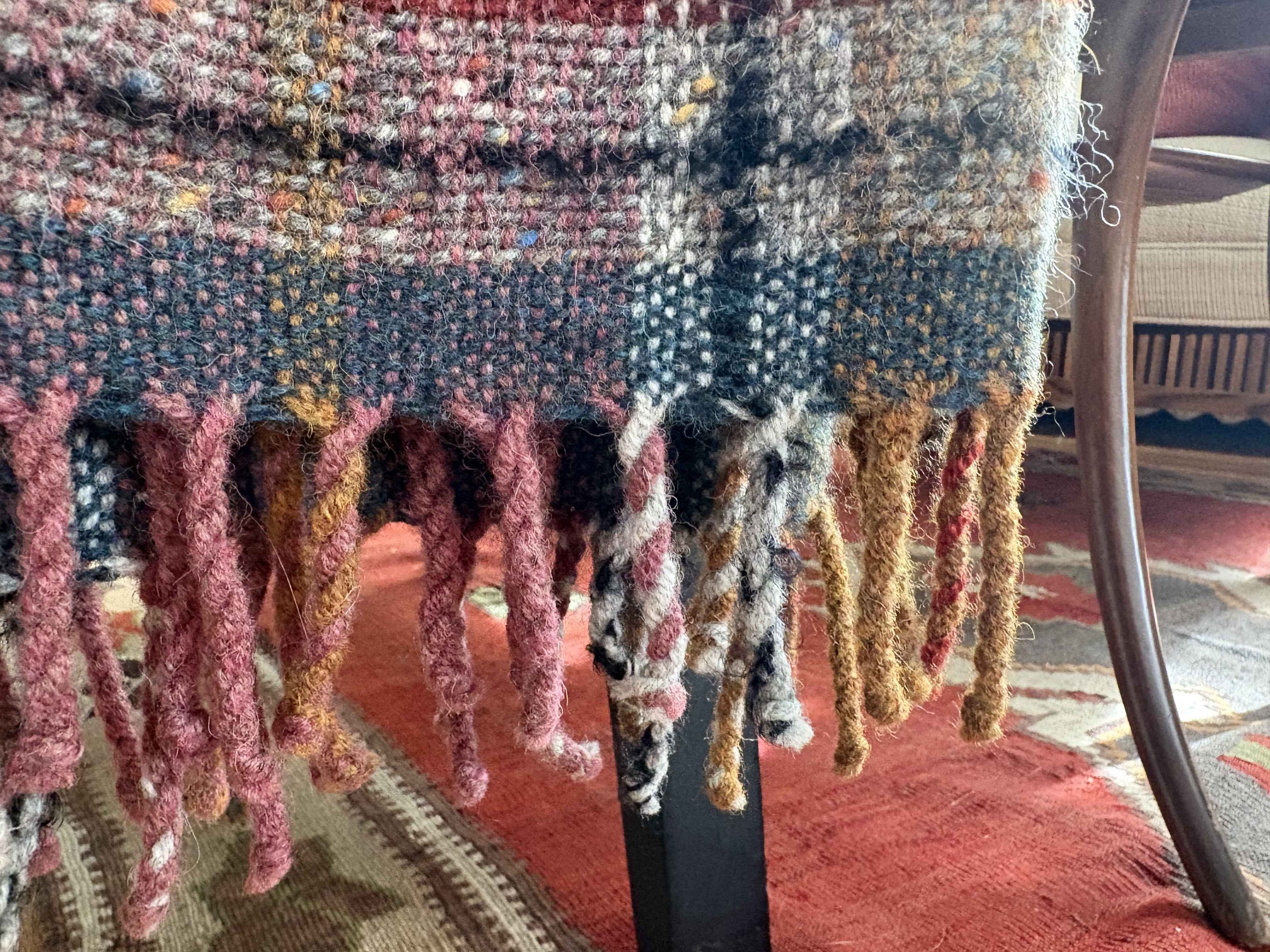 Close-up of a woven textile with colorful yarns, hanging from a support structure, with a red carpet and wooden furniture visible in the background.