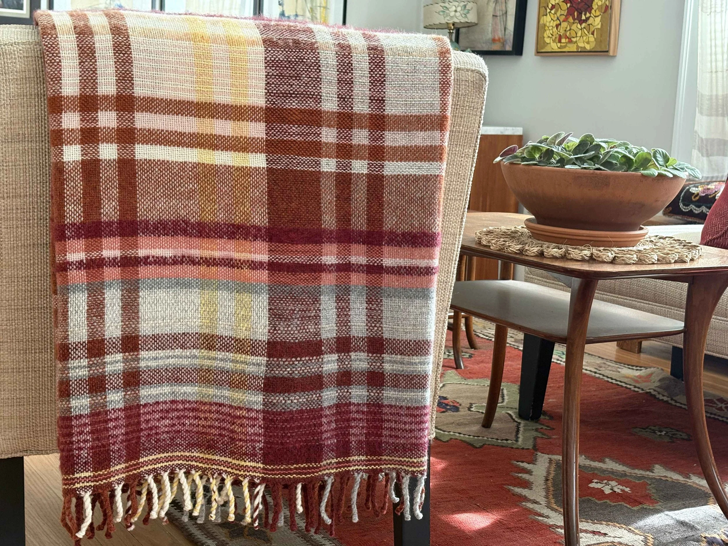 A beige upholstered armchair with a red and white checked plaid throw blanket hanging over its back, next to a wooden side table with a potted plant on it, in a room decorated with colorful artwork on the walls and a patterned rug.