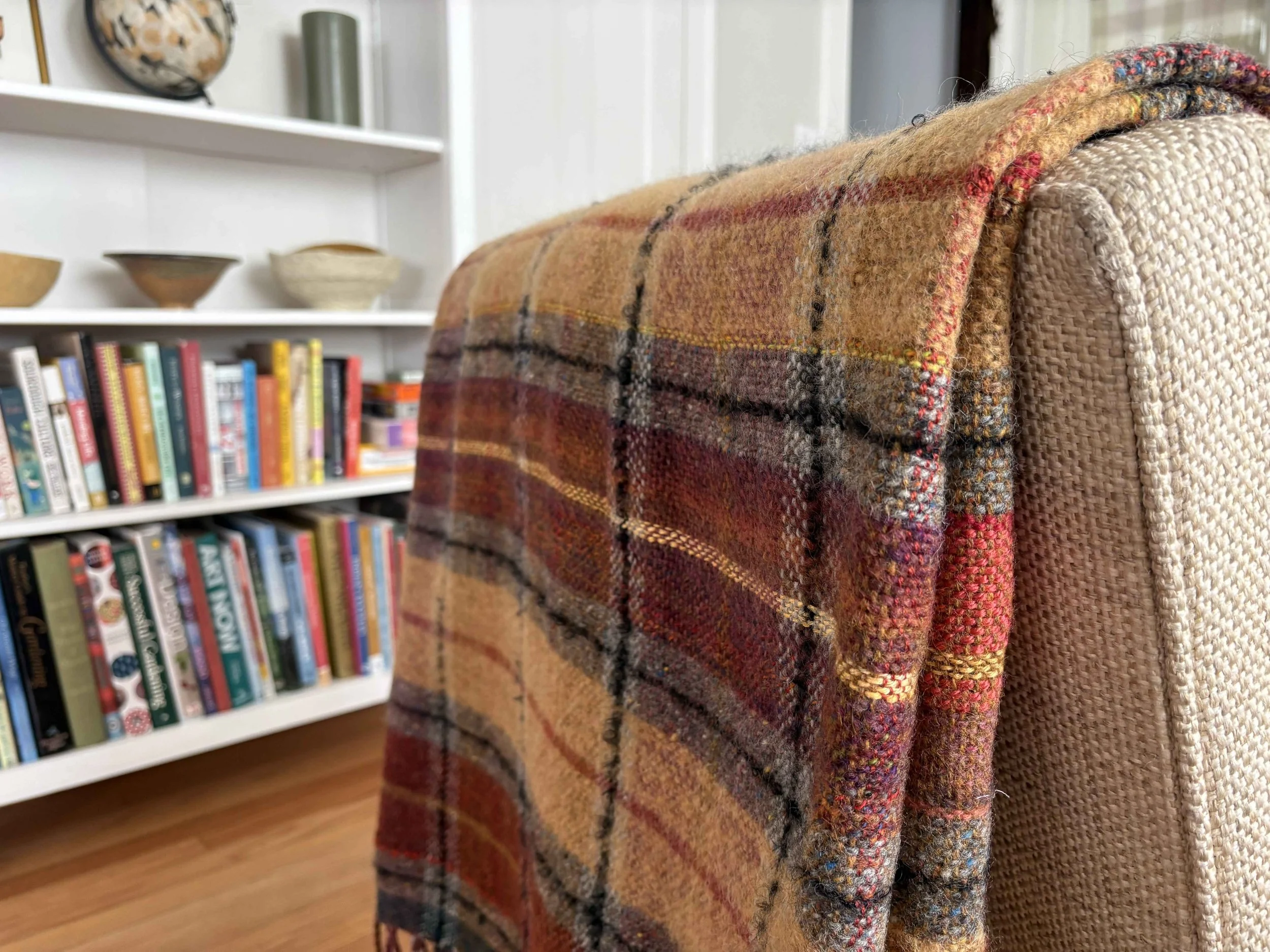 Close-up view of a woven, patterned blanket draped over the back of a textured beige armchair, with a bookshelf filled with books and decorative bowls in the background.