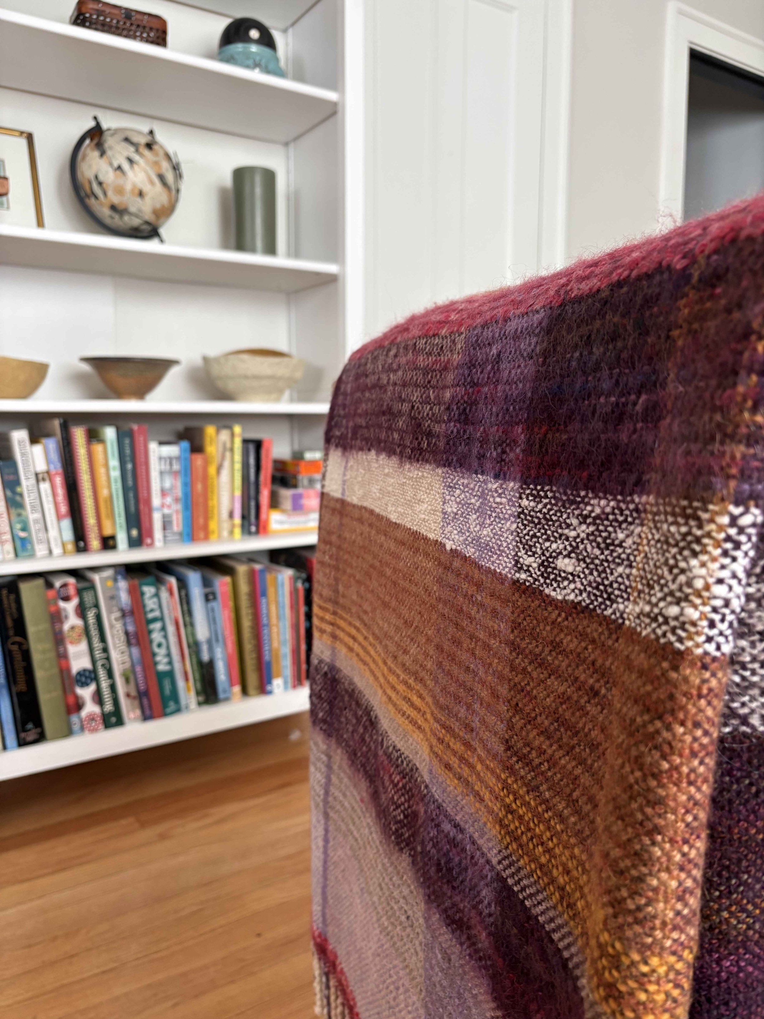 Close-up of a colorful woven fabric on the back of a chair in a room with a bookshelf and white walls.