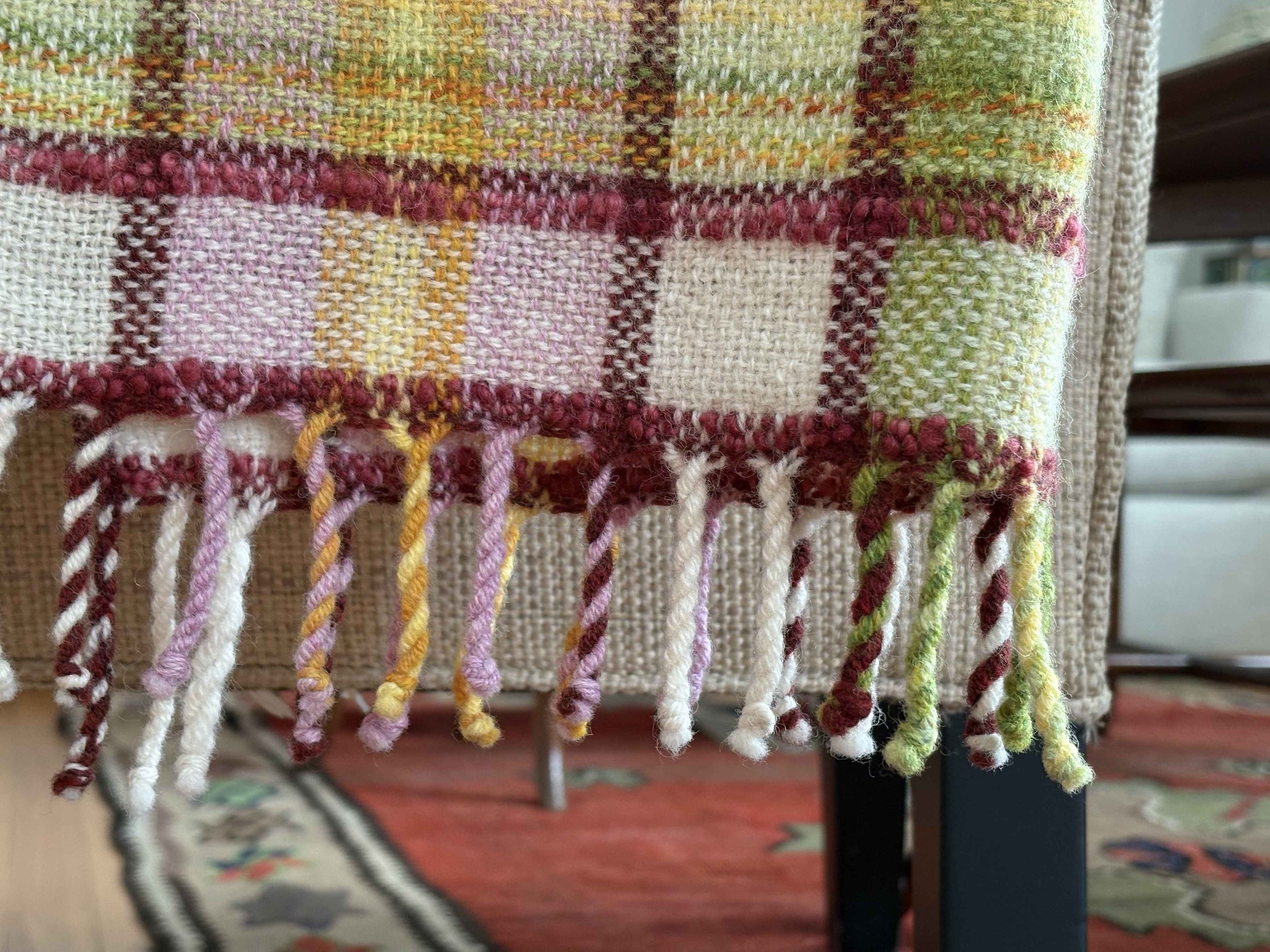 Close-up of colorful woven fabric with fringe, featuring checkered pattern in shades of pink, red, yellow, green, and white, hanging from a piece of furniture.