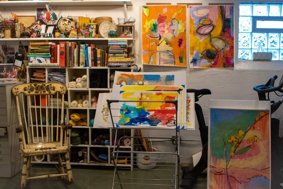 An art studio with colorful abstract paintings on the wall, a bookshelf filled with books and craft supplies, a vintage wooden chair, and a metal art easel holding a colorful painting. There are yarn skeins stored in cubbies and a window with blue-tinted glass letting in natural light.