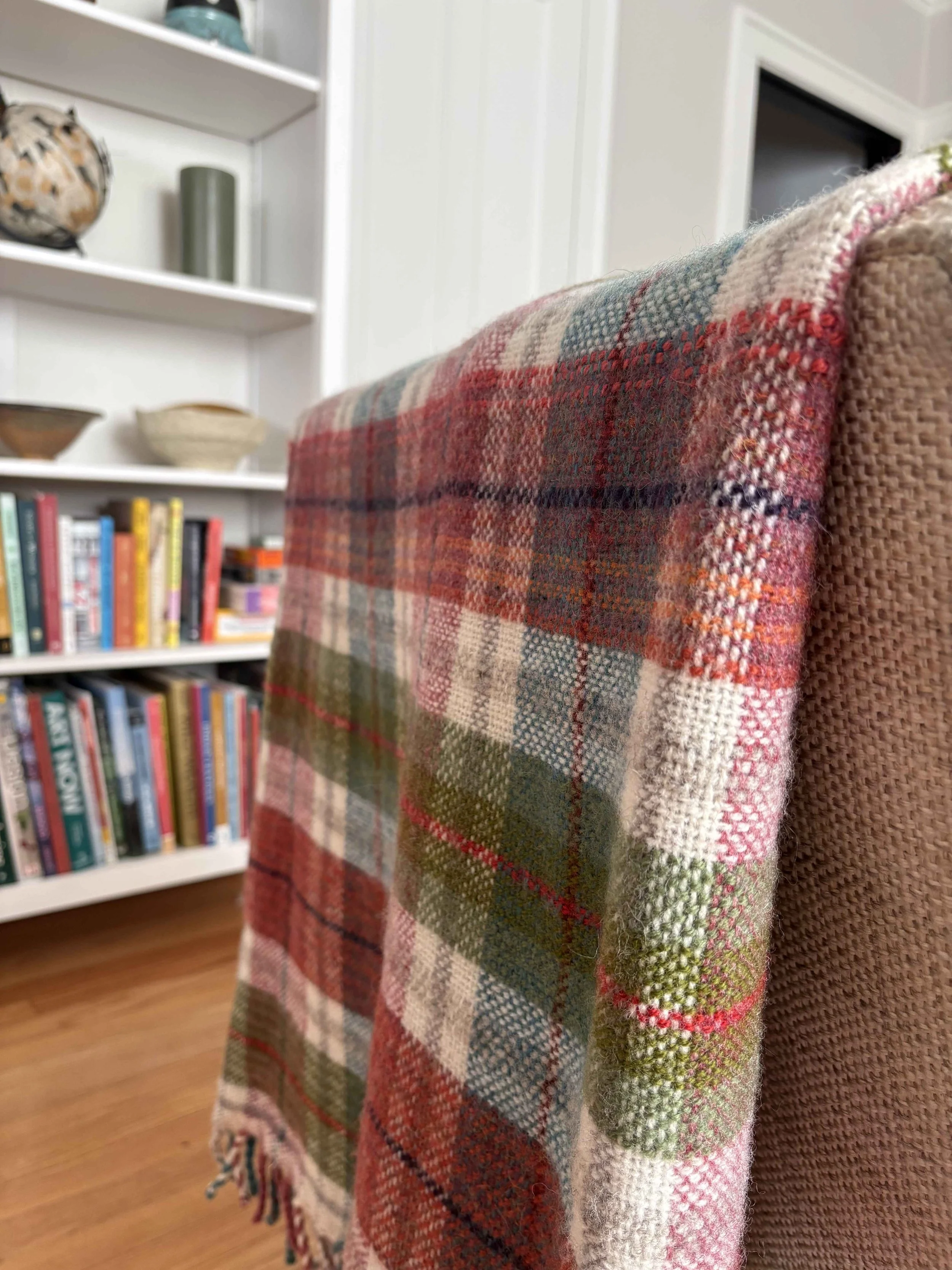 Close-up of a plaid blanket draped over the back of a pink upholstered chair in a room with a white bookshelf filled with books, decorative bowls, and vases in the background.