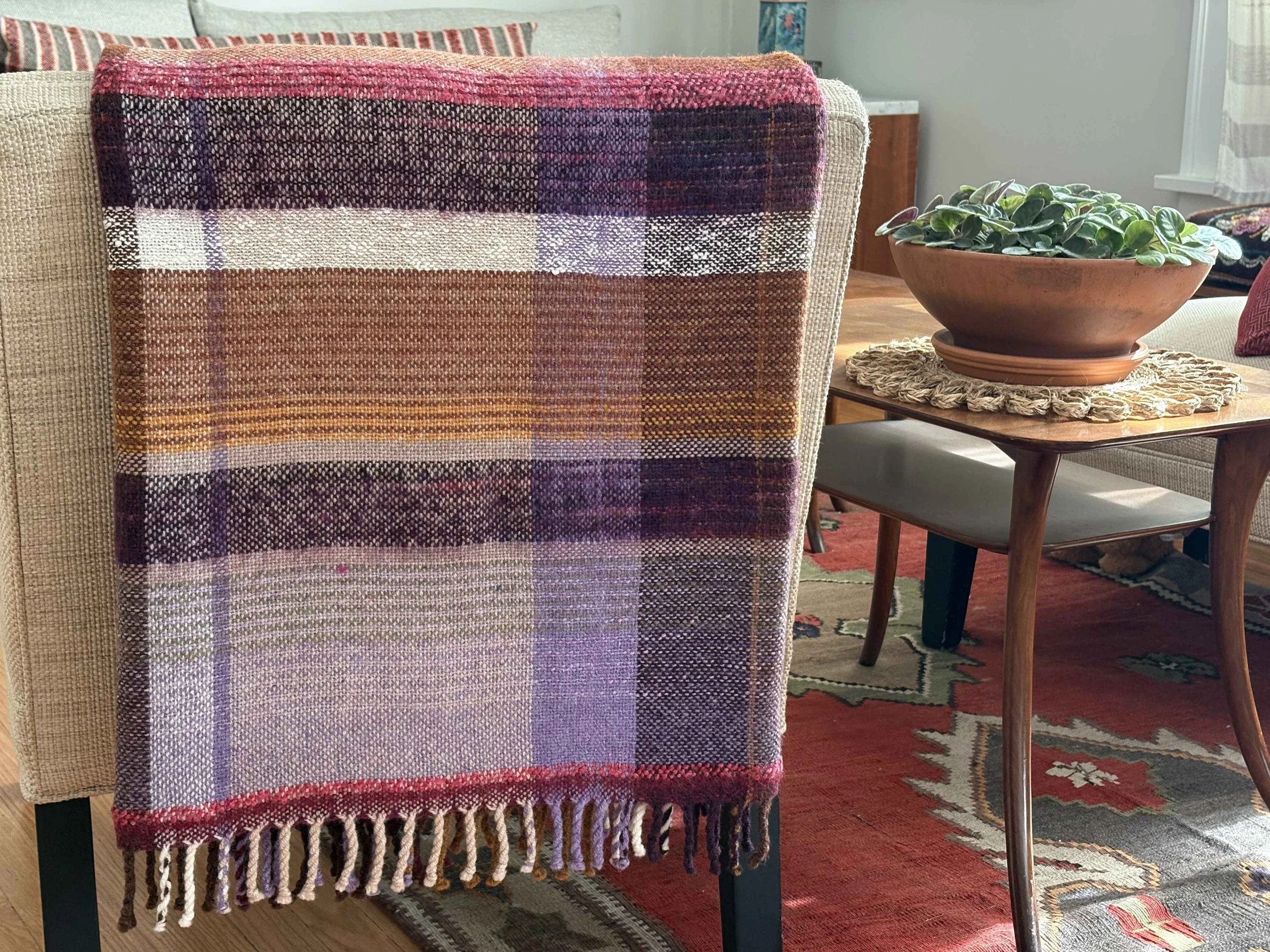 A close-up of a colorful woven textile with fringes hanging over a chair arm, beside a wooden table with a round woven placemat and a potted plant.