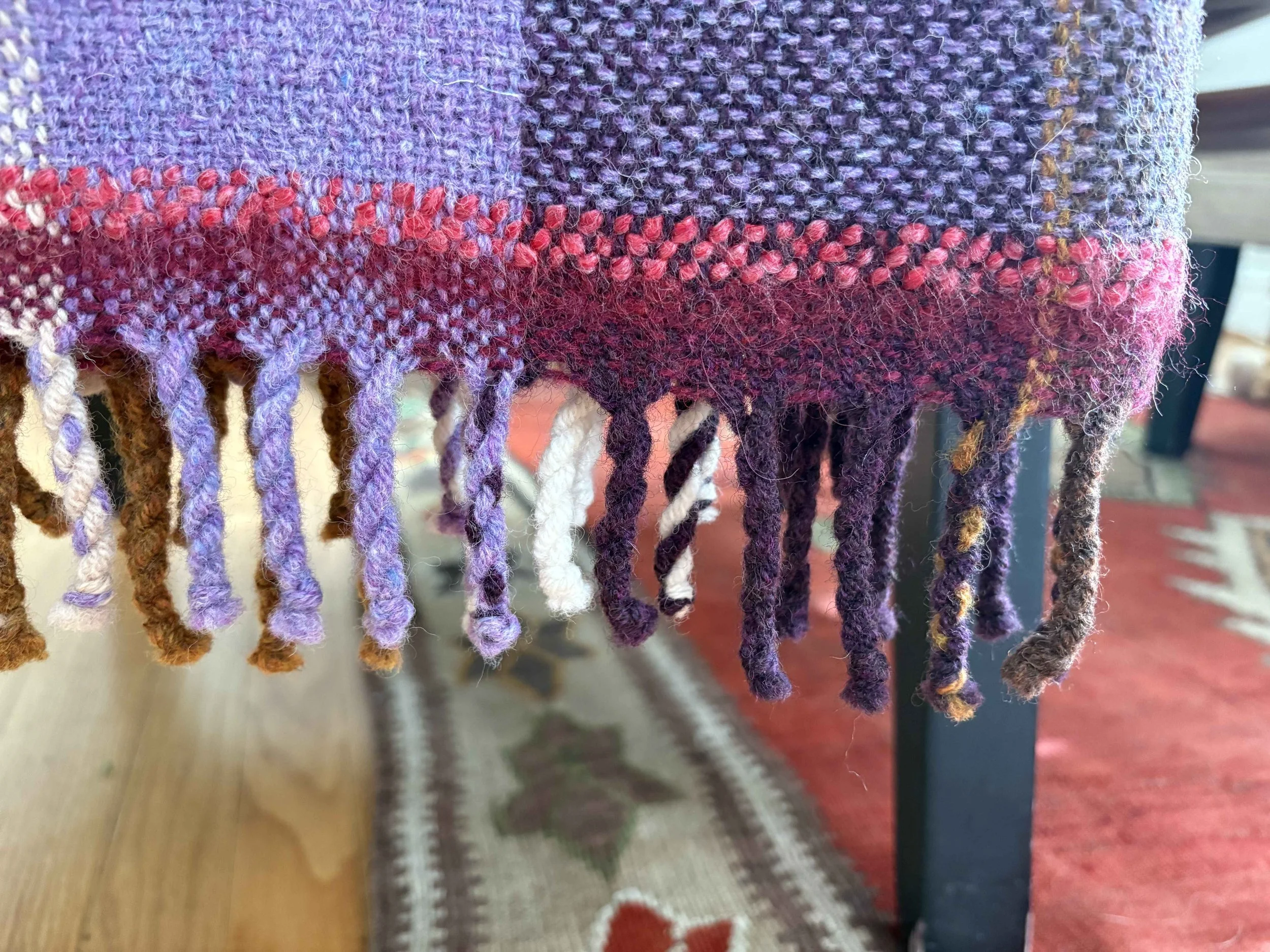 Close-up of a woven fabric with purple, red, pink, and multicolored strands, and fringes hanging from the edge.