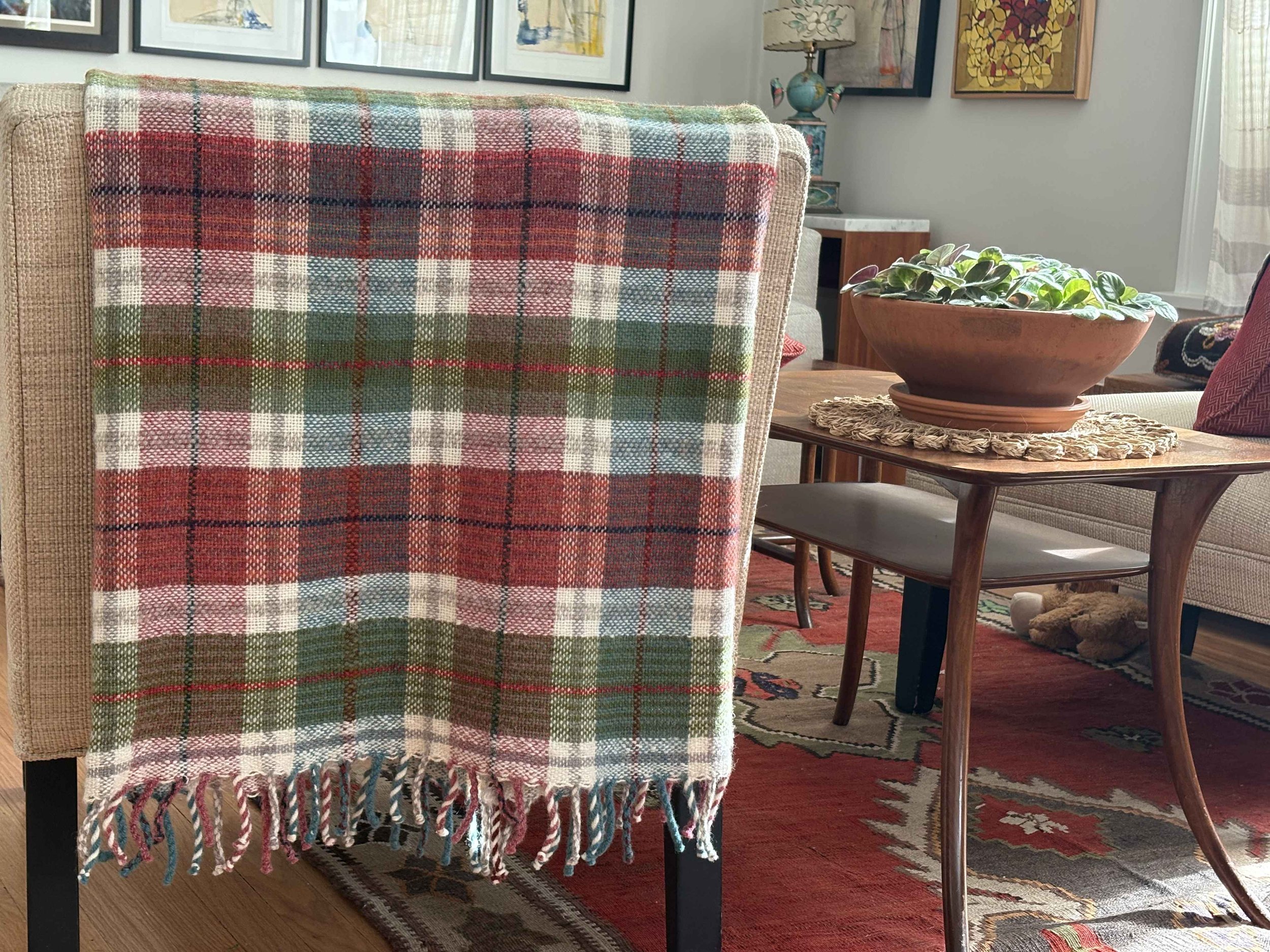 A dining room with a beige upholstered chair covered with a red, green, and cream plaid throw blanket. Behind, a collection of framed artwork hangs on the wall. To the right, a small table with a potted plant in a large terracotta bowl and a woven doily underneath. Part of a red and black patterned rug is visible on the hardwood floor.