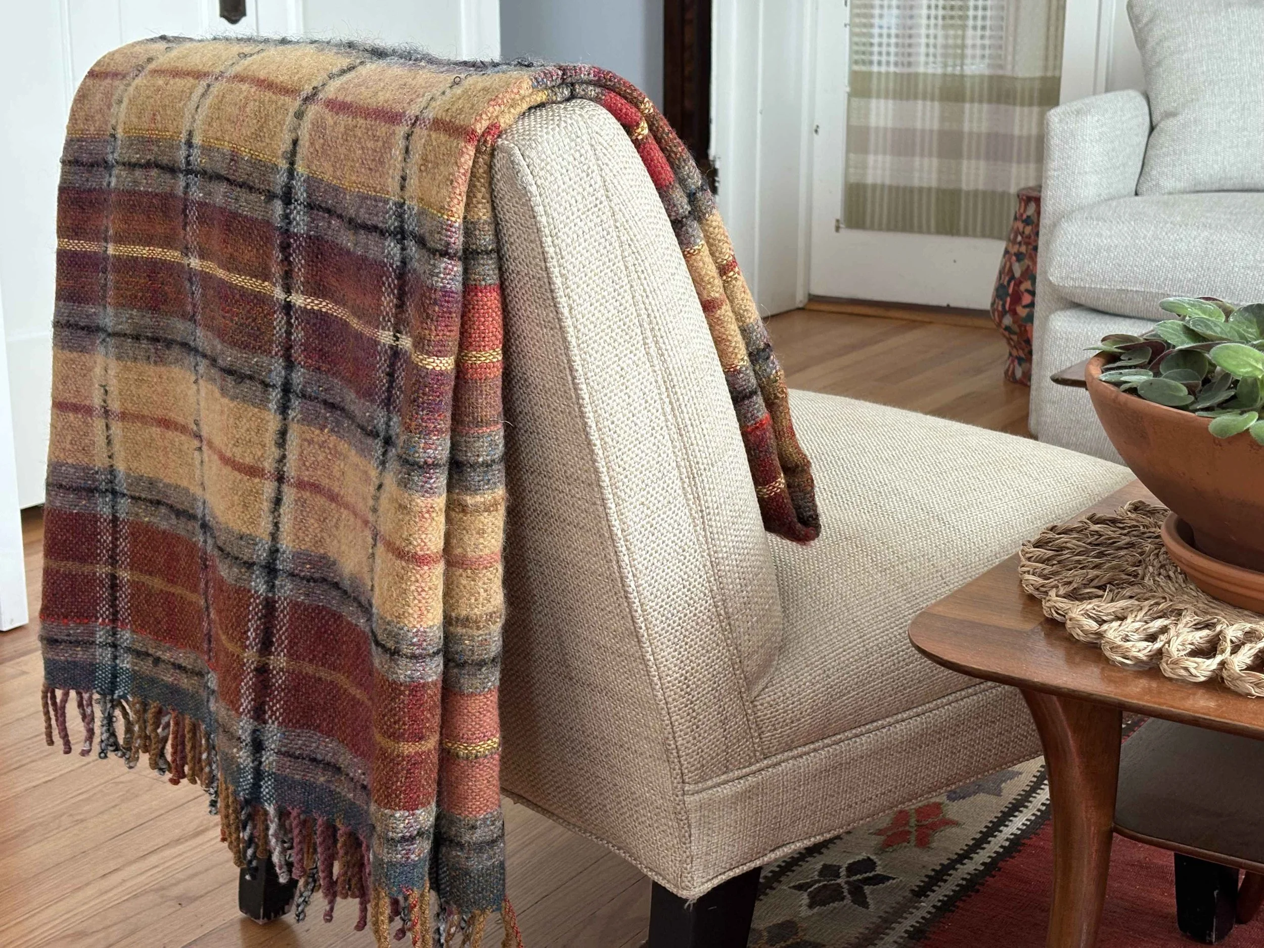 A beige upholstered armchair with a multicolored plaid woven blanket draped over its back. To the right, a wooden side table with a woven basket and a potted plant. In the background, a window with green striped curtains and a closed white door.