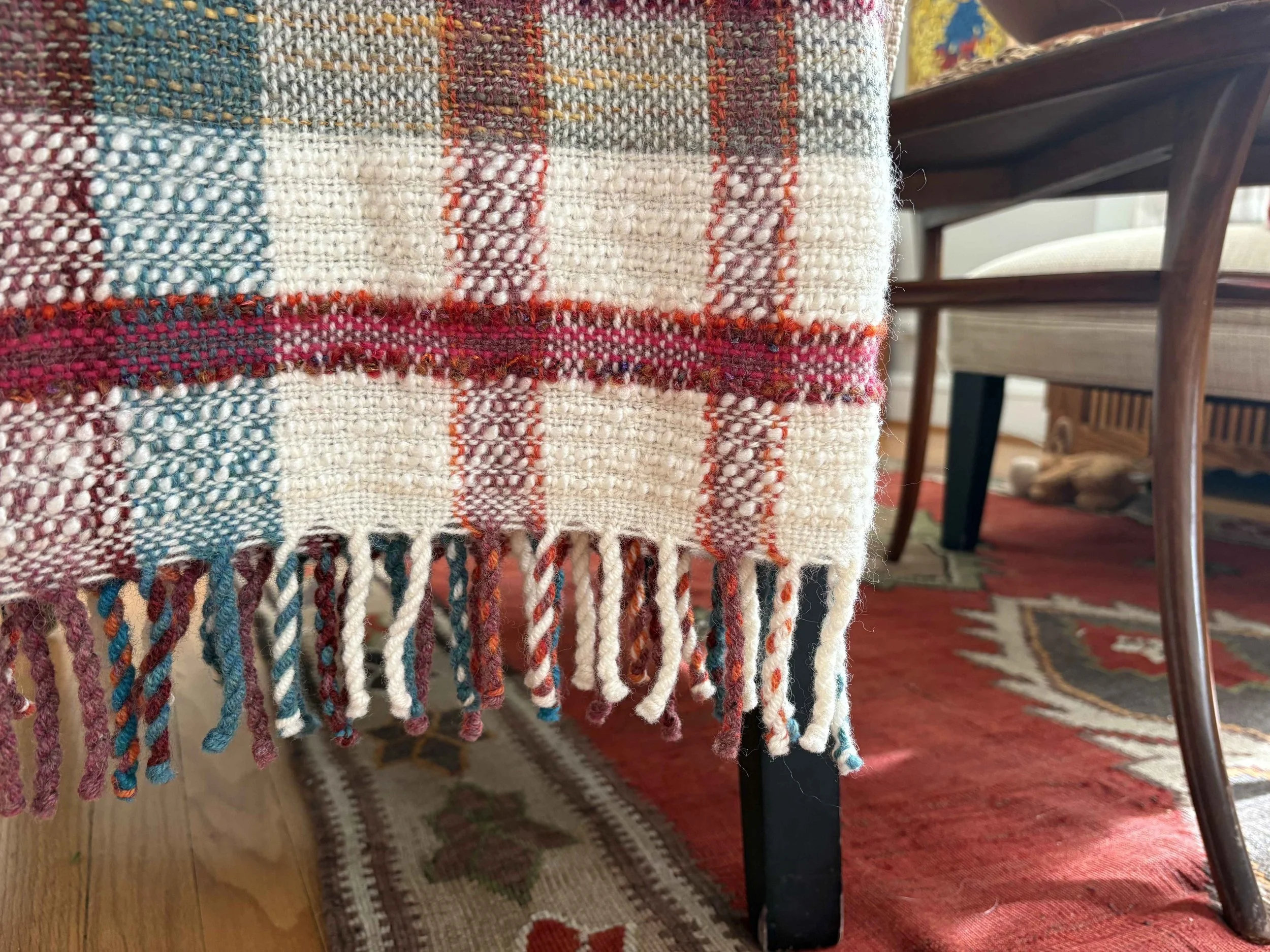 Close-up of a woven fabric with colorful plaid pattern and fringed edge, on a piece of furniture in a room with wooden flooring, a patterned rug, and chairs.