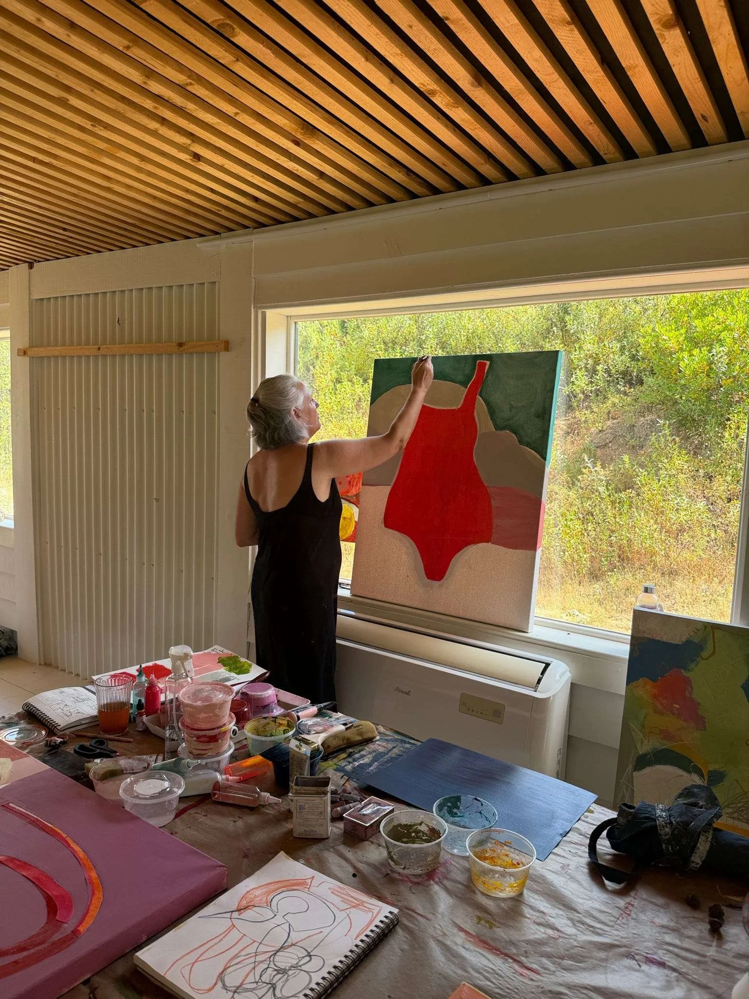 An older woman with gray hair painting a red dress on a canvas in her art studio, with various art supplies and drawings on the table, and a large window showing greenery outside.