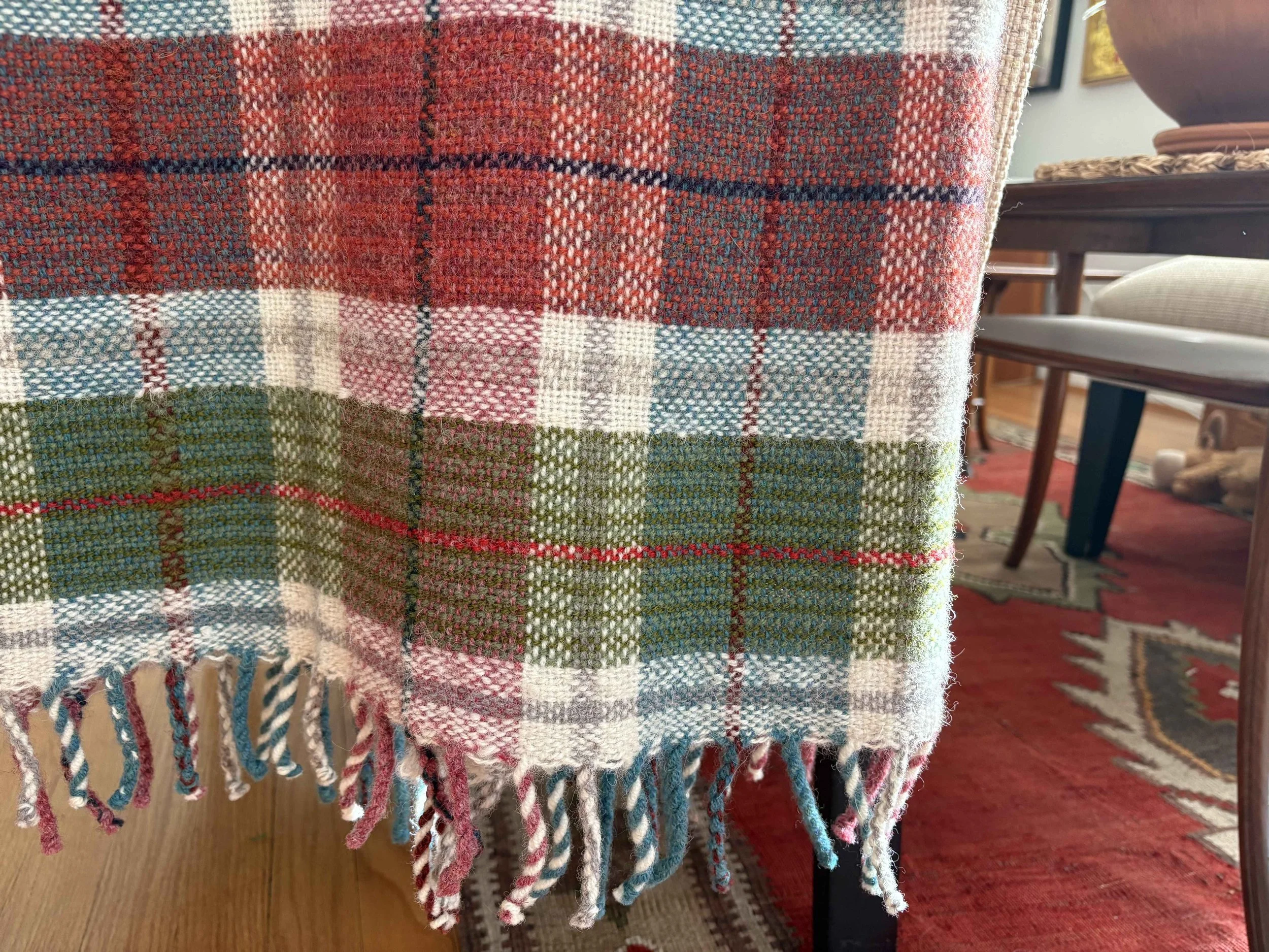 Close-up of a colorful plaid woven blanket with fringed edges, hanging over a piece of furniture in a living room.