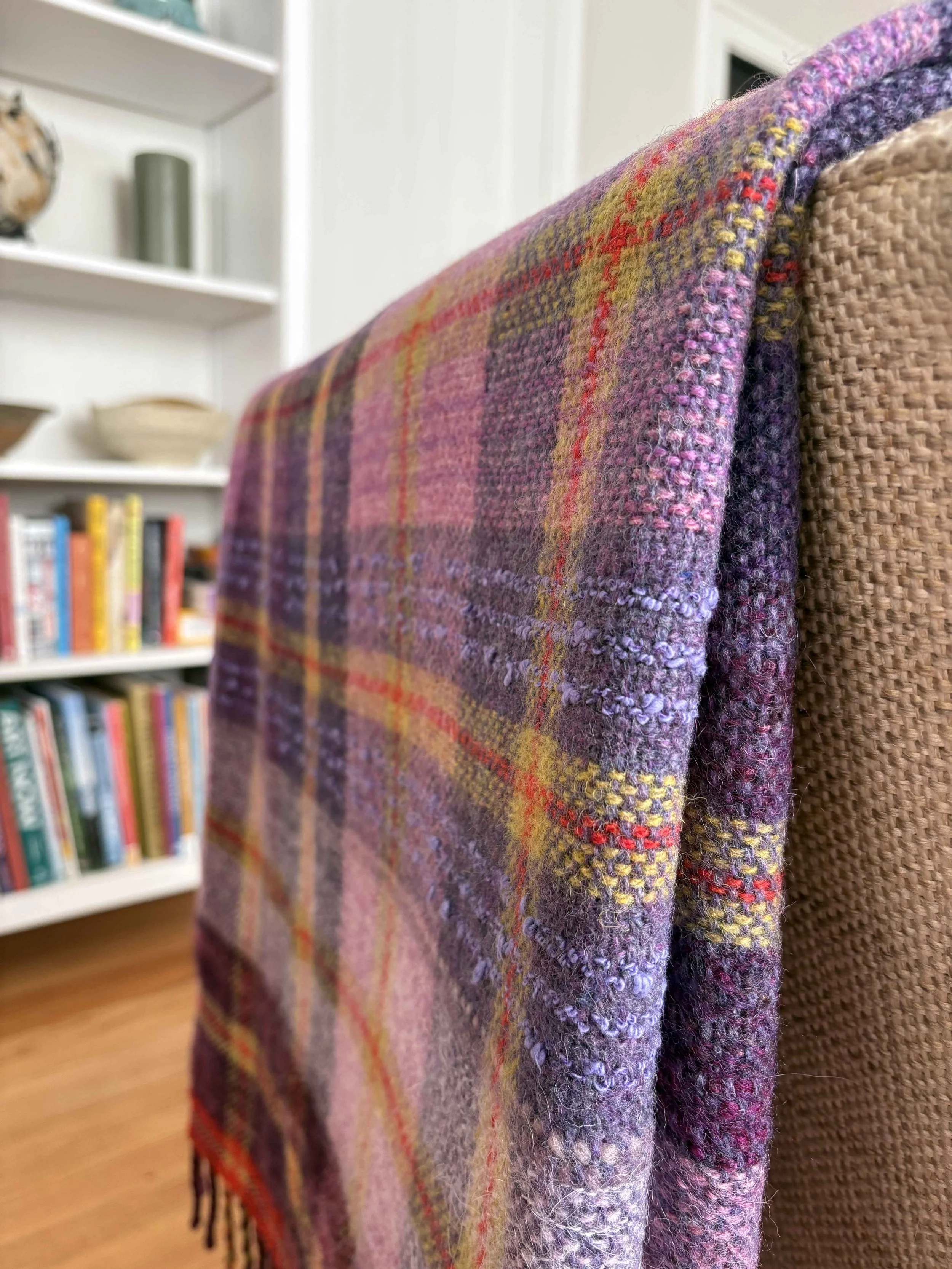 Close-up of a woven, plaid wool fabric with shades of purple, pink, and red draped over the back of a woven chair, with a bookshelf in the background.