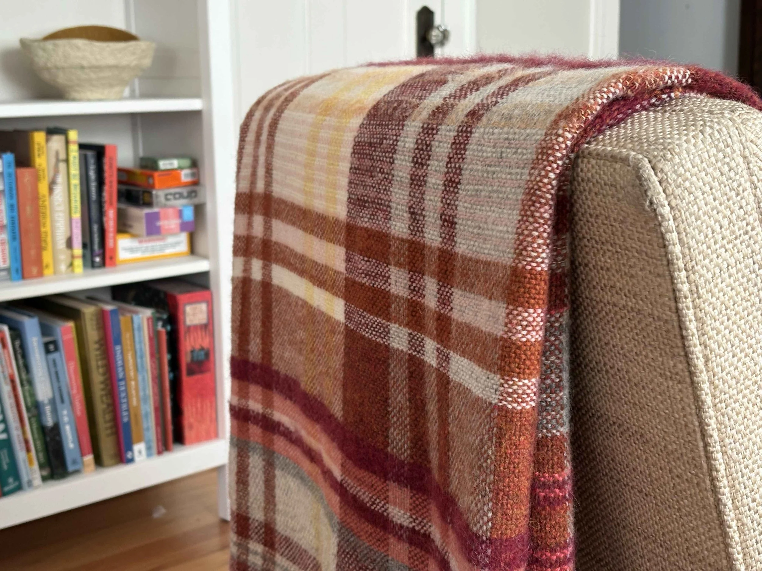 Close-up of a colorful plaid blanket draped over the arm of an upholstered chair in front of a bookshelf filled with books.