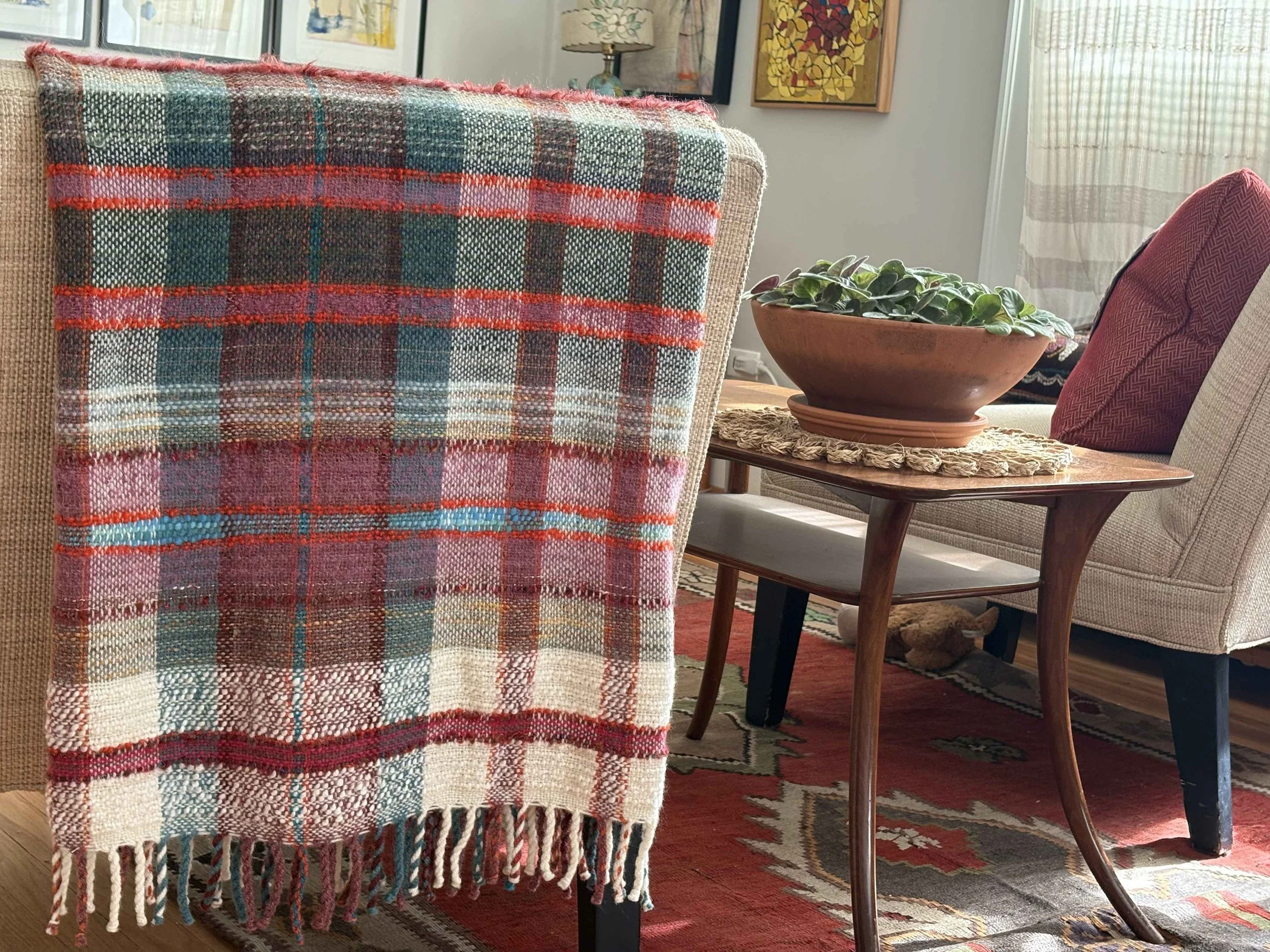 Close-up of a plaid woven blanket with red, green, pink, and beige colors draped over the back of a beige armchair in a cozy living room setting. Behind the chair, there is a wooden side table with a potted plant, and an interior decorated with frame