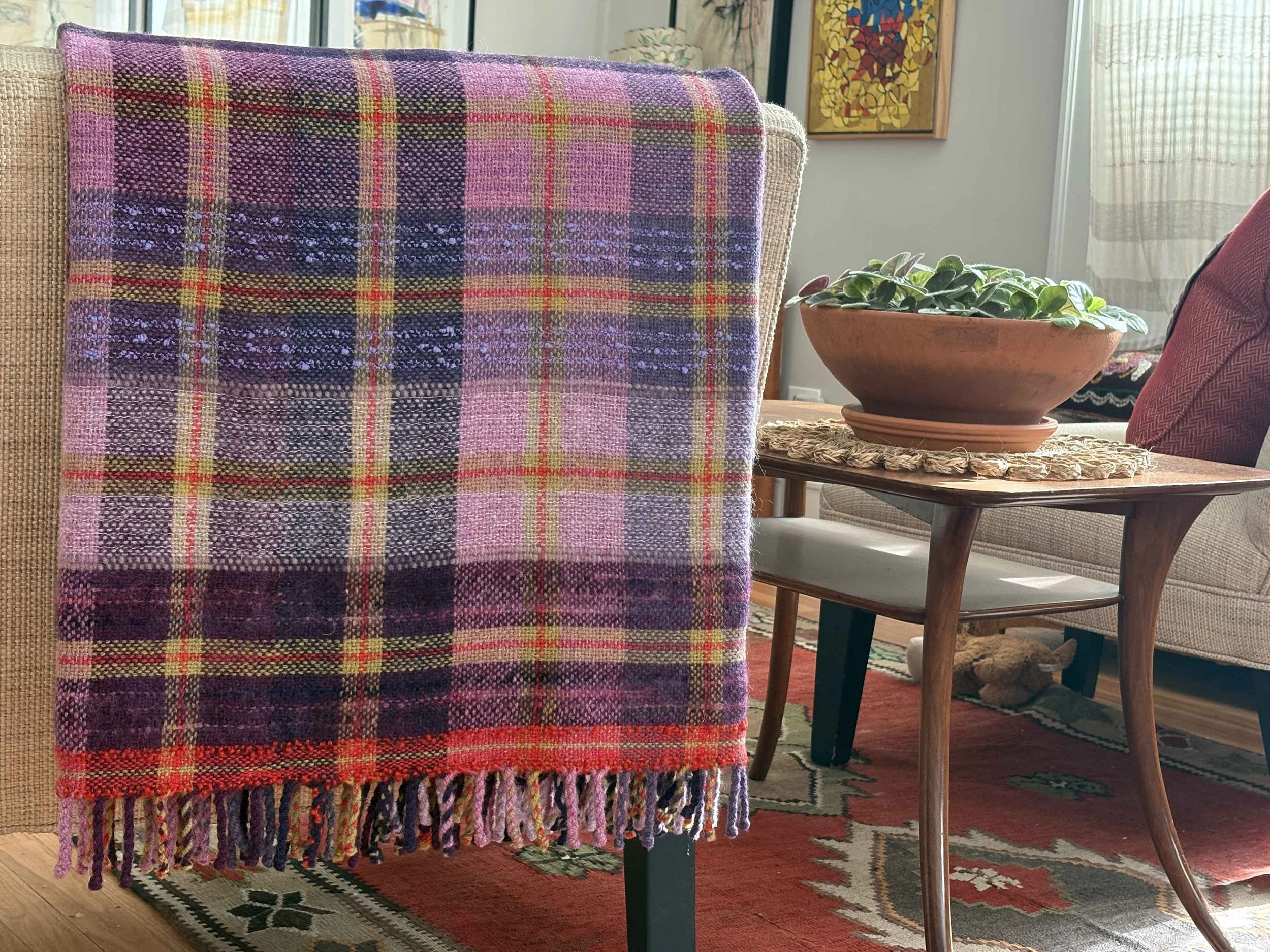 A plaid blanket with pink, purple, yellow, black, and red colors hanging over the back of a beige upholstered chair, in a cozy living room setting.