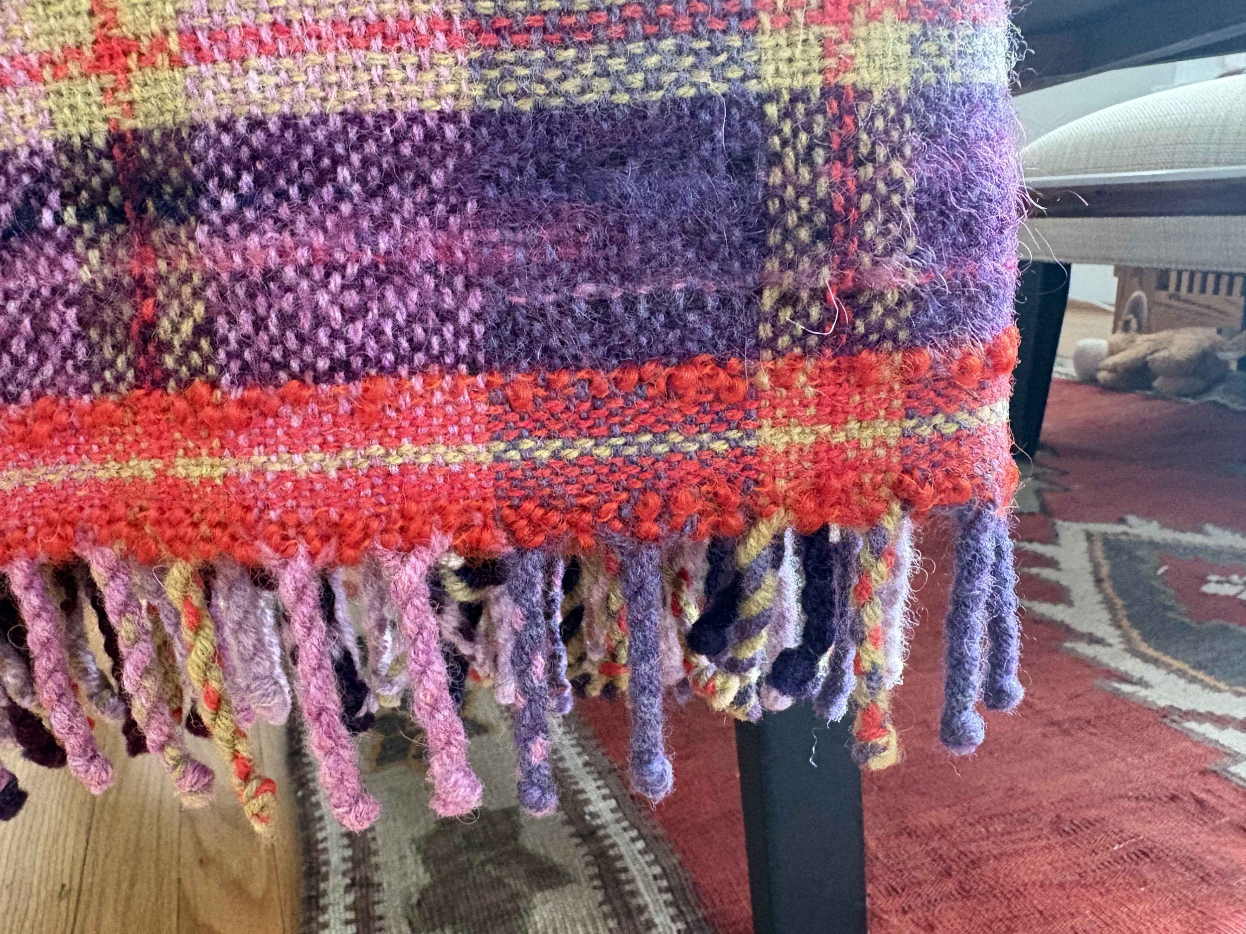 Close-up view of a colorful, textured, knitted or woven fabric with fringed edges, featuring various shades of purple, pink, red, and yellow. Part of a patterned rug and furniture are visible in the background.