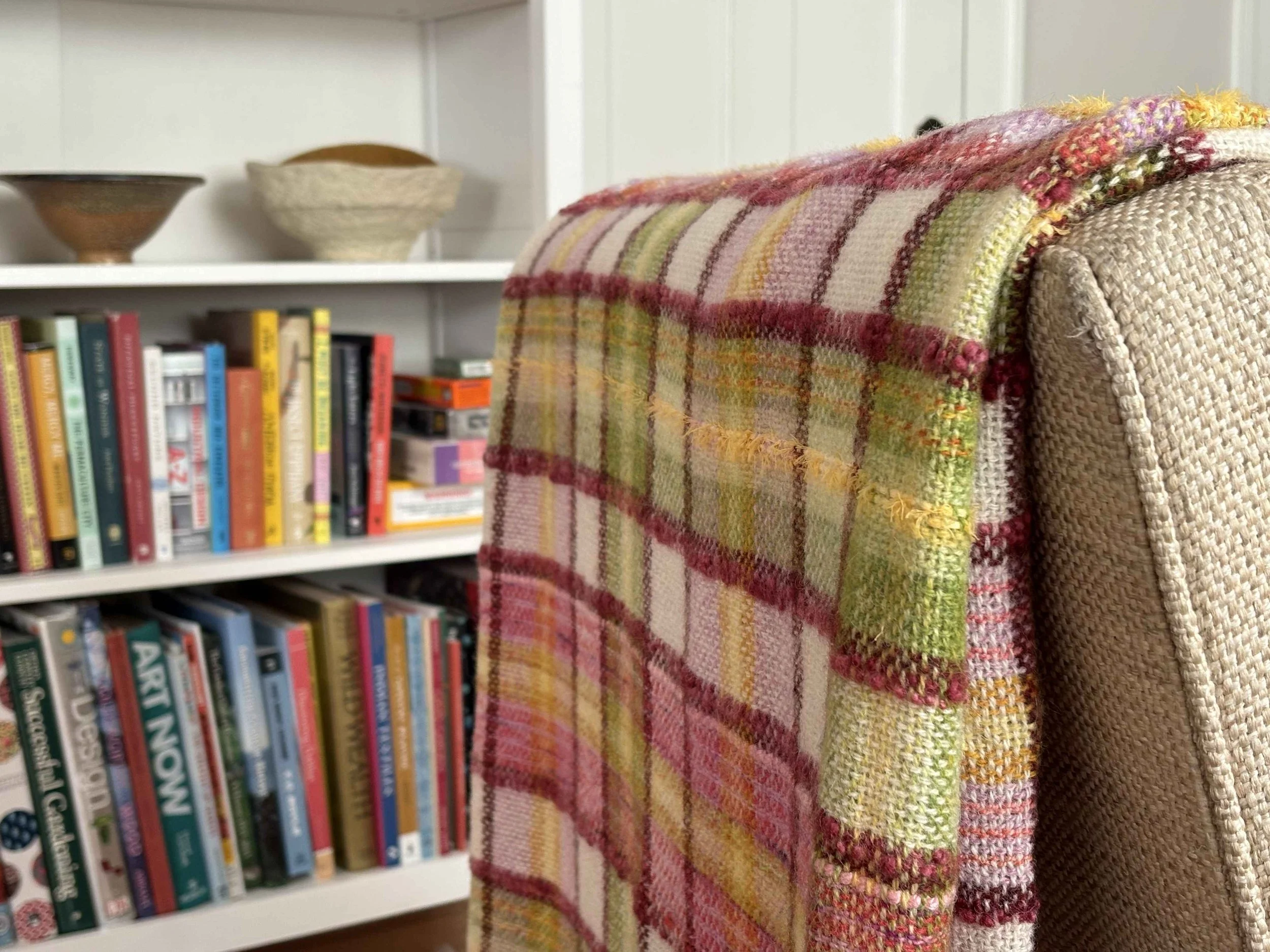 Colorful plaid blanket draped over the back of a beige woven armchair in a room with white walls, a bookshelf filled with various books, and two decorative bowls on a white shelf.