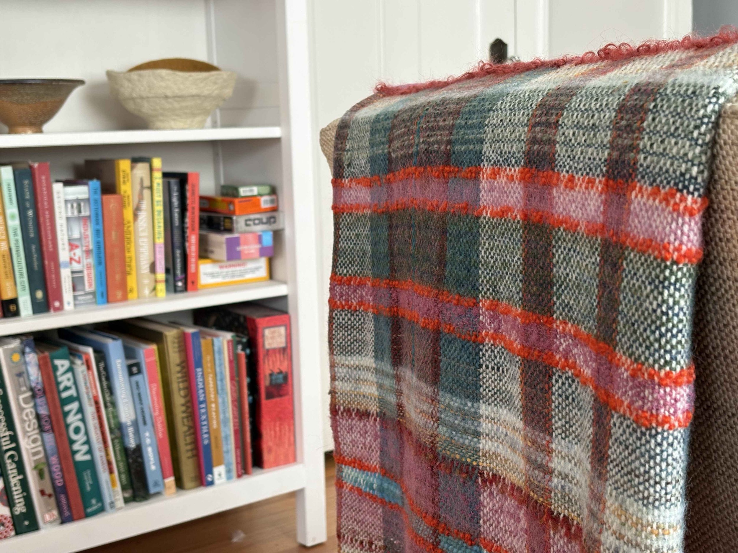 Close-up of a colorful, patterned woven textile hanging on a chair in front of a white bookshelf filled with books and boxes.