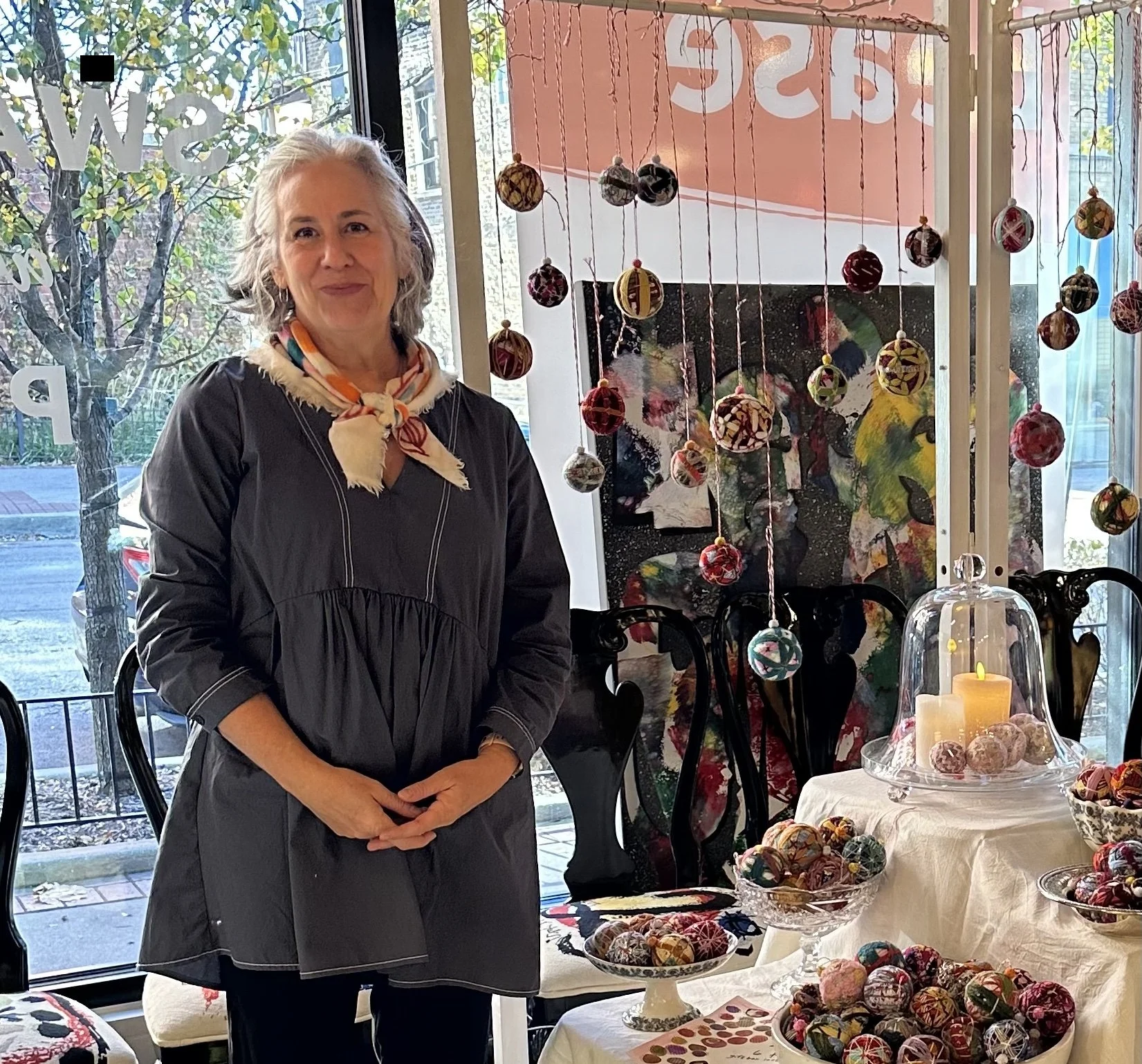 A woman with gray hair standing inside a shop, surrounded by colorful fabric-covered balls displayed on tables and hanging decorations. Outside the window, trees and a street are visible.