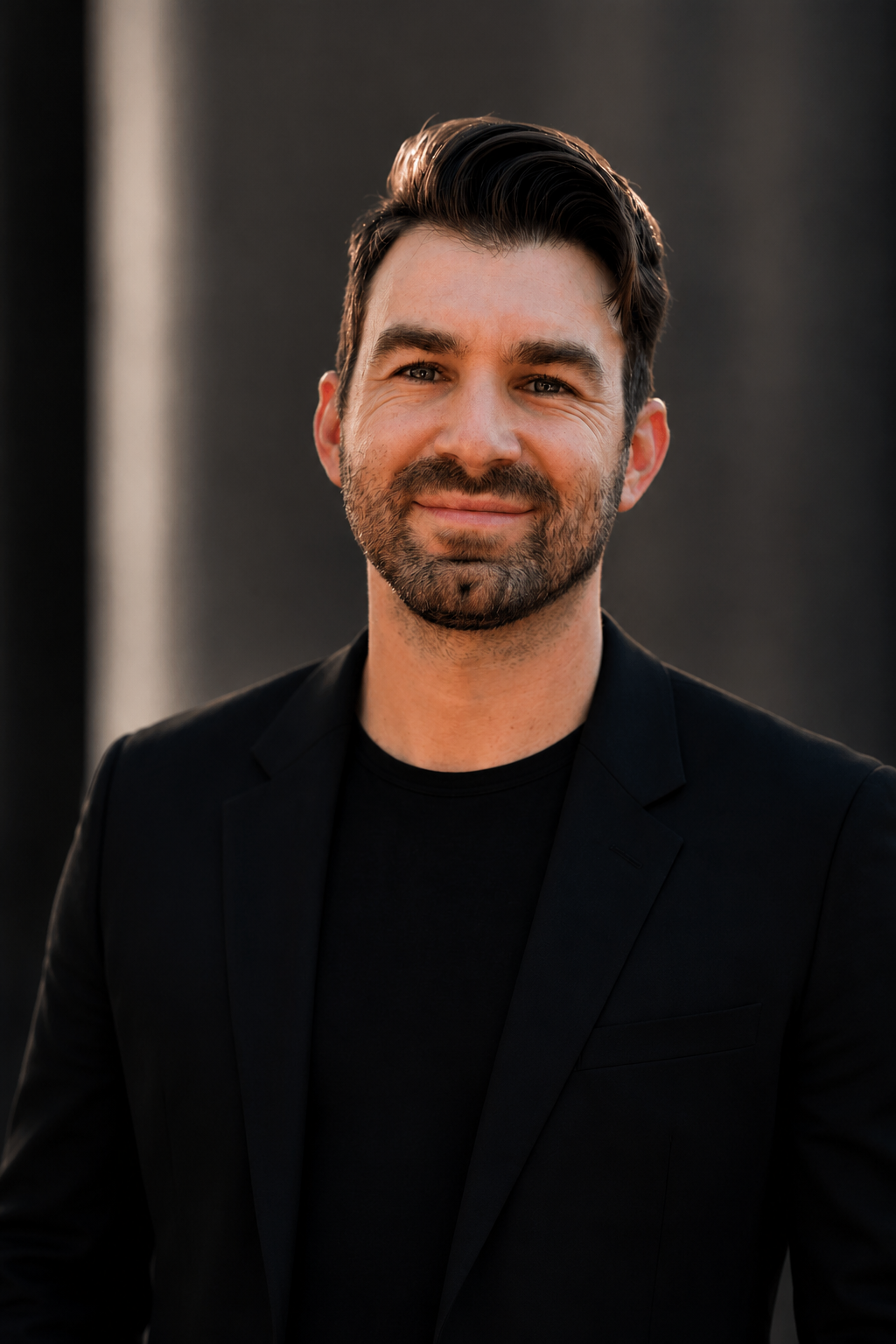 A smiling man with dark hair and a beard wearing a black blazer and shirt, standing outdoors with a dark blurred background.